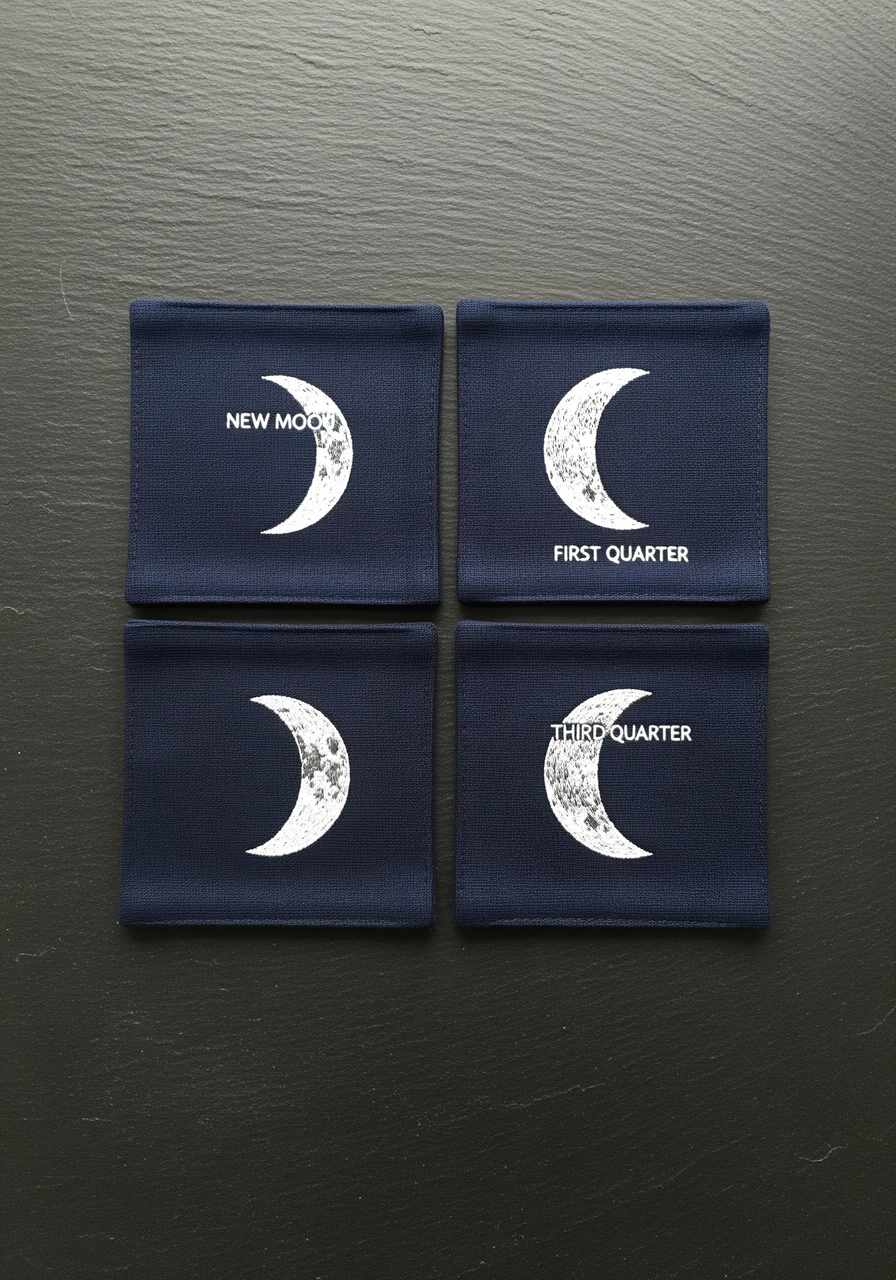 Four navy blue coasters, each embroidered with a different phase of the moon in white.