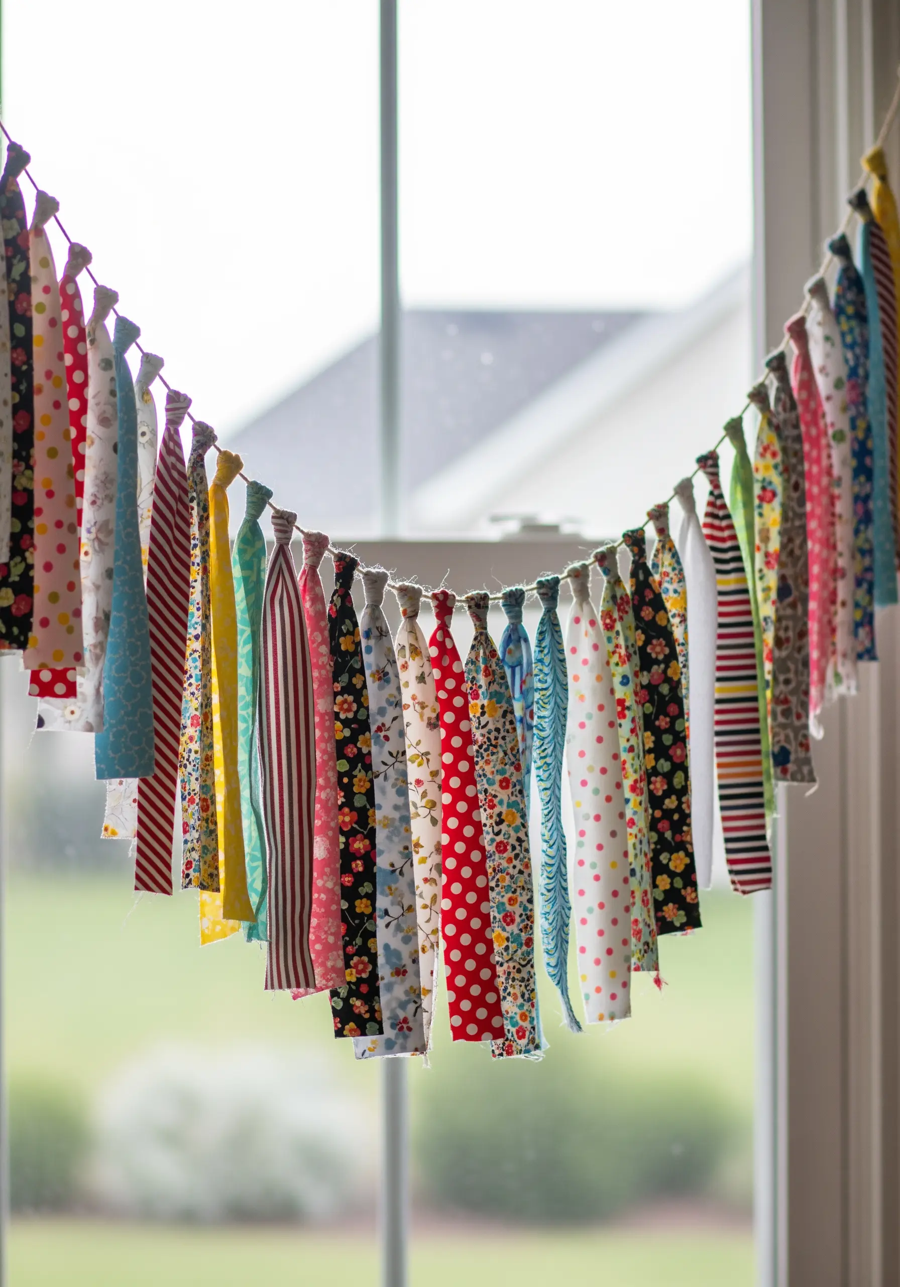 A colorful garland made of knotted fabric scraps hanging in front of a window.