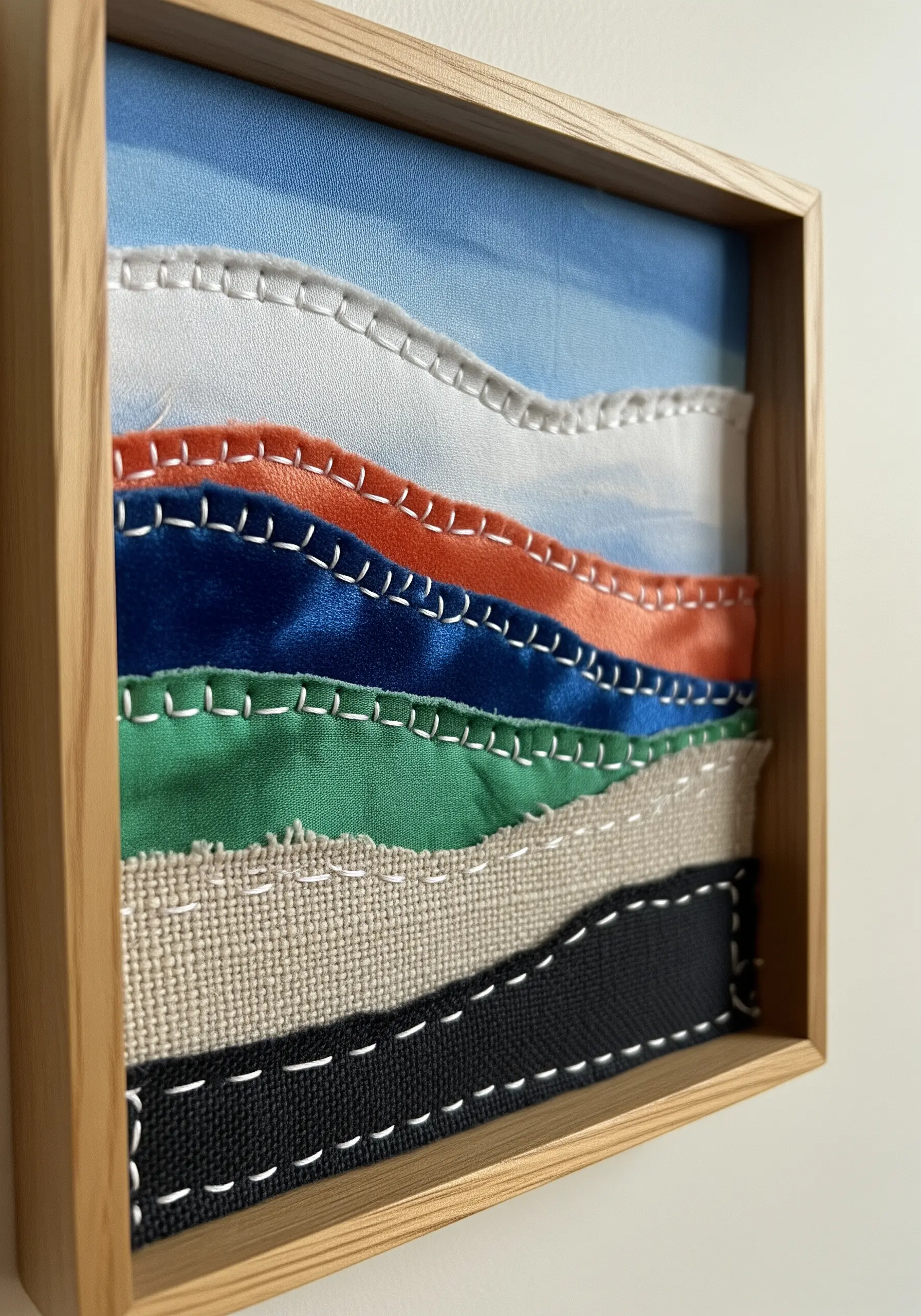 Framed fabric collage landscape with layers of colorful textiles and visible white stitching.