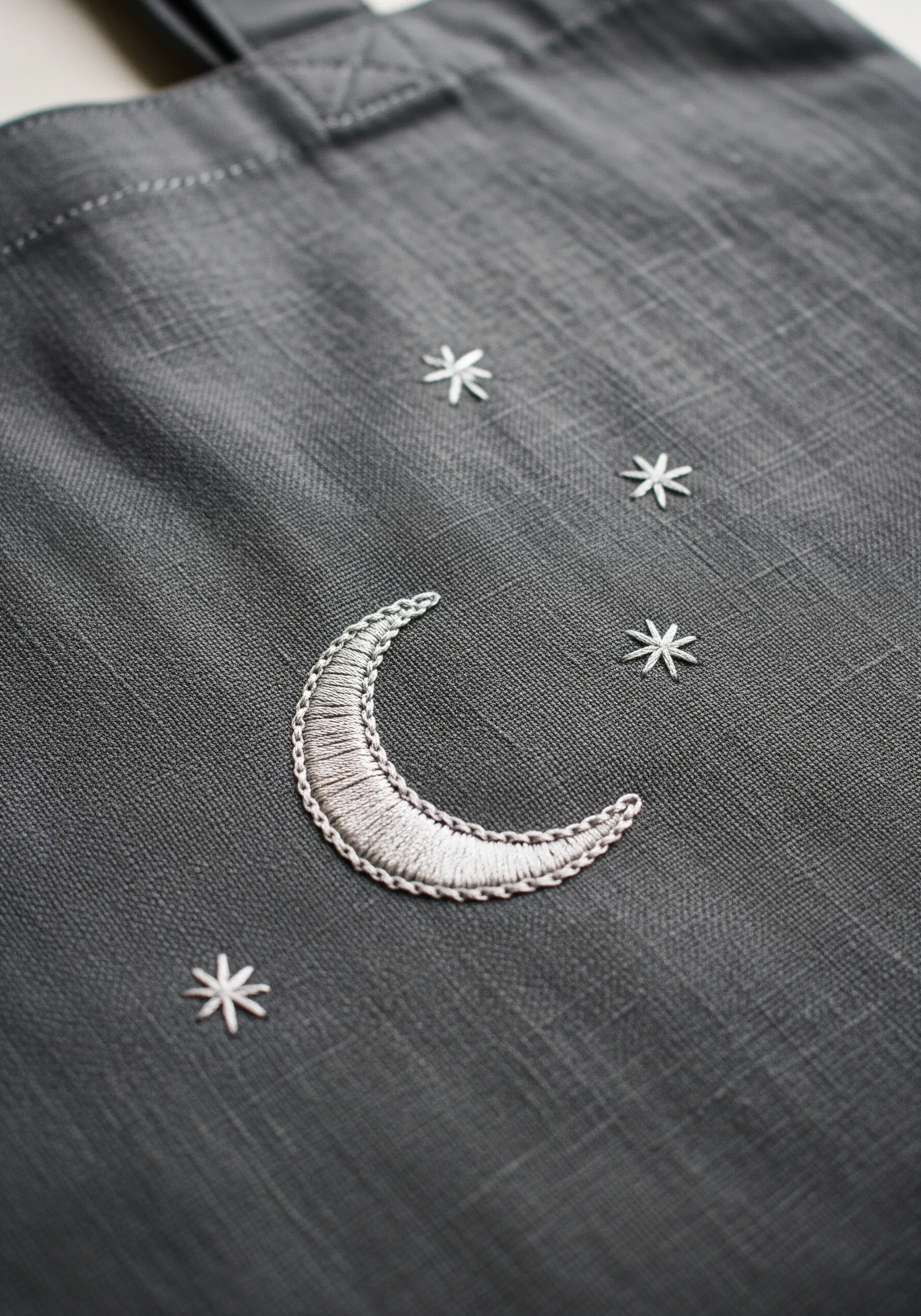 A padded satin stitch crescent moon in silver thread on a dark gray tote bag with small stars.