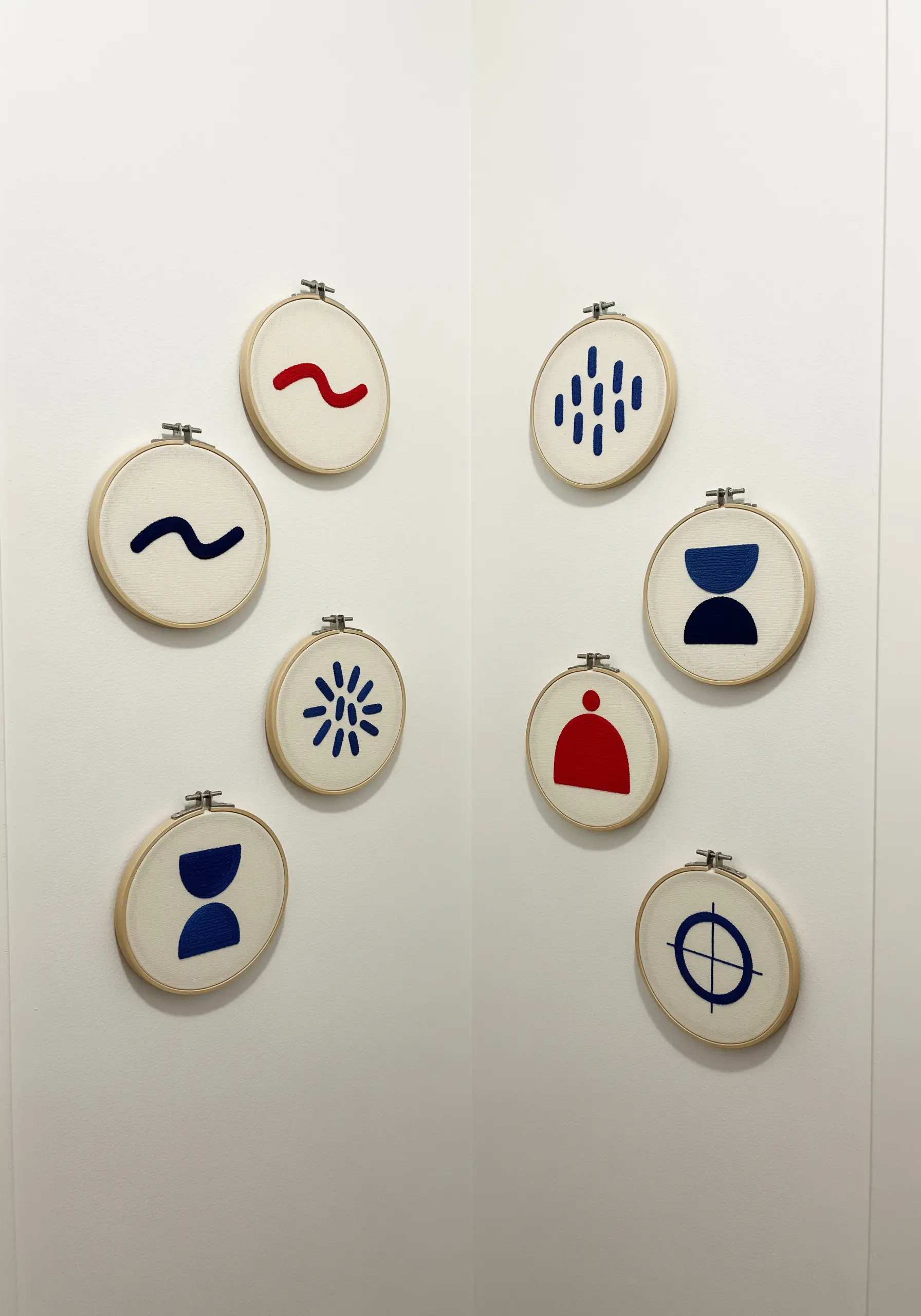 A scattered collection of eight small embroidery hoops, each with a simple abstract symbol in red or blue.