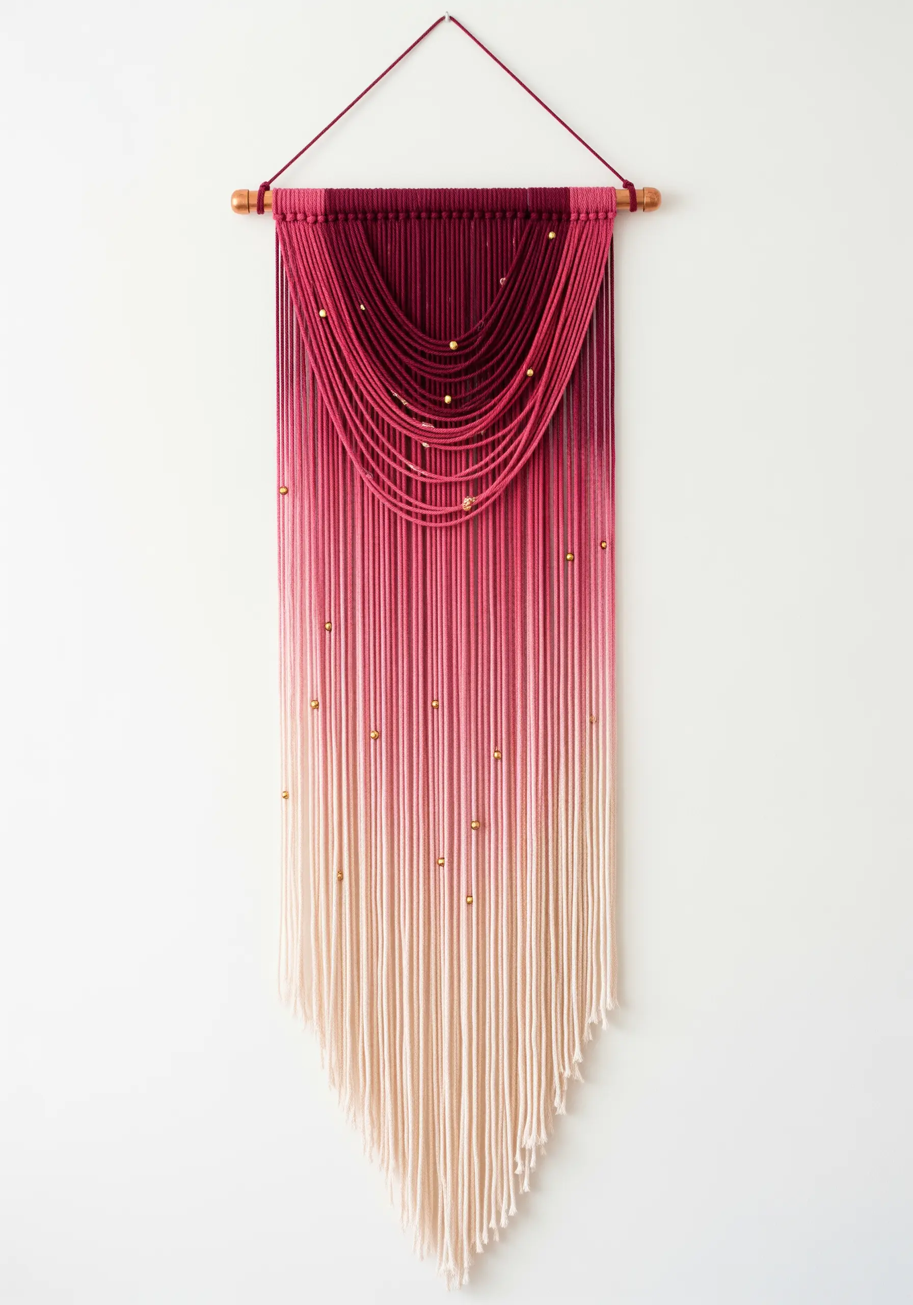 A dip-dyed wall hanging with a color gradient from deep magenta to pale pink, accented with gold beads.
