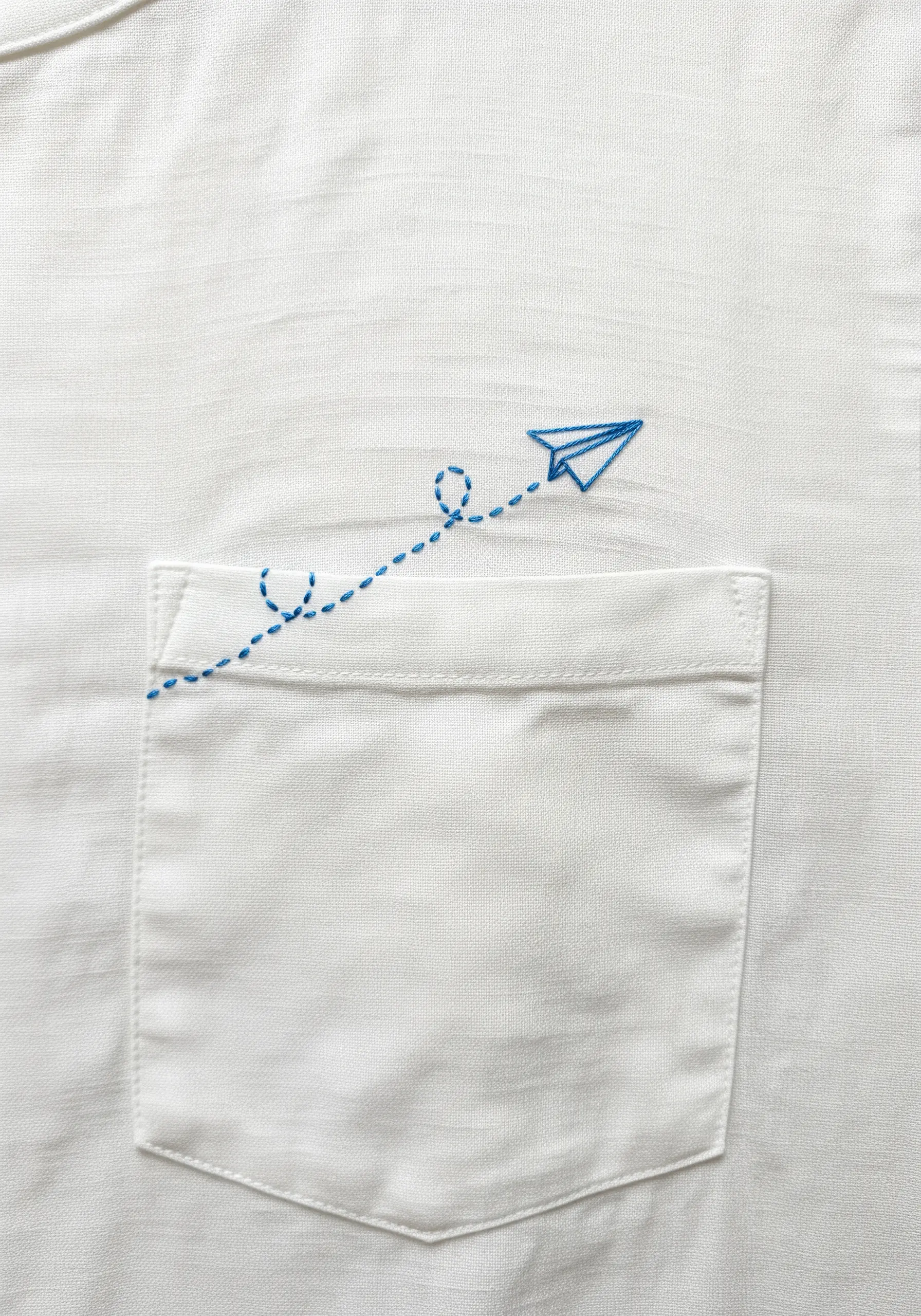 A small blue paper airplane embroidered above a shirt pocket with a dashed-line flight path.
