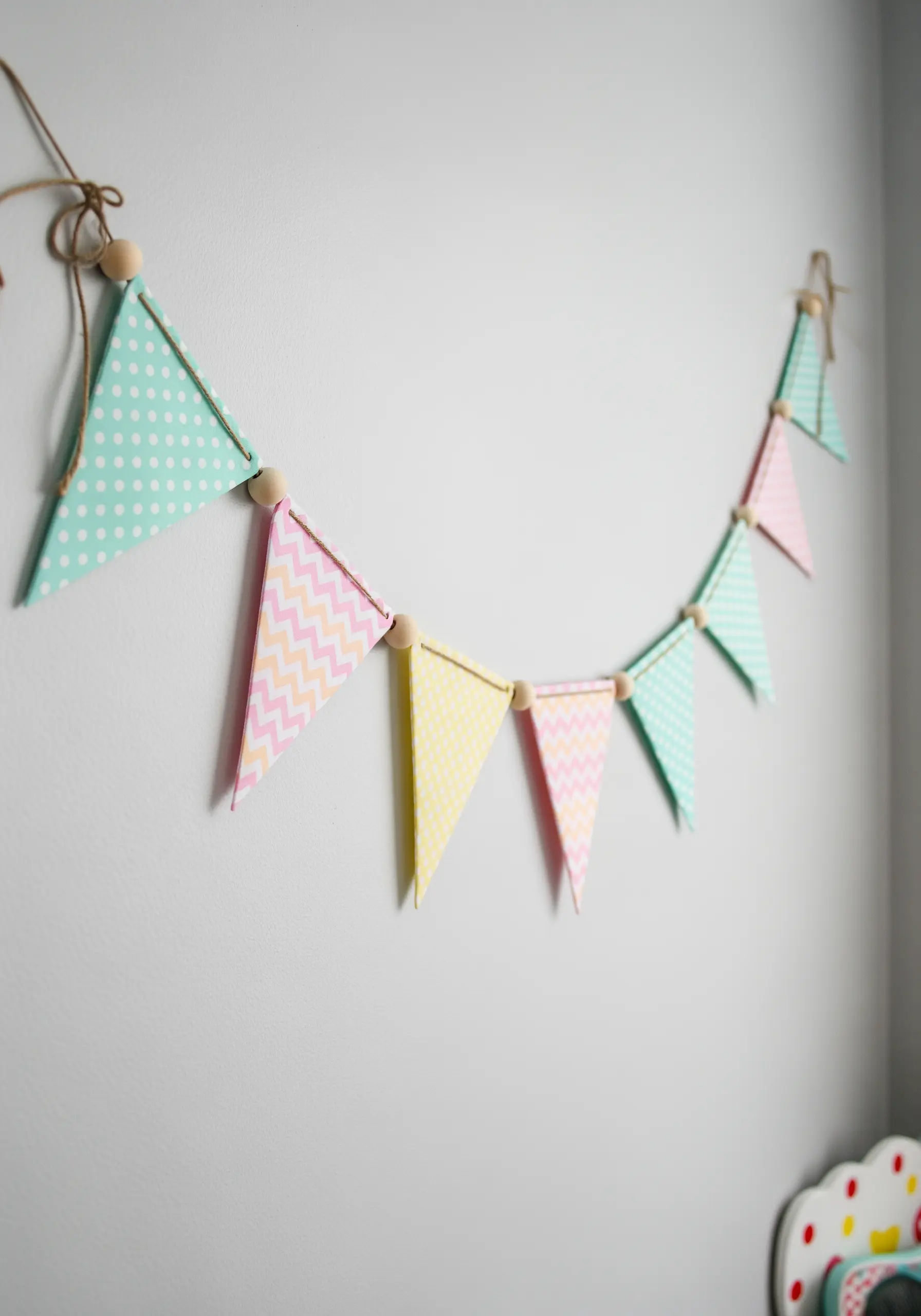 A pastel-colored fabric bunting with wooden beads hanging on a white wall.