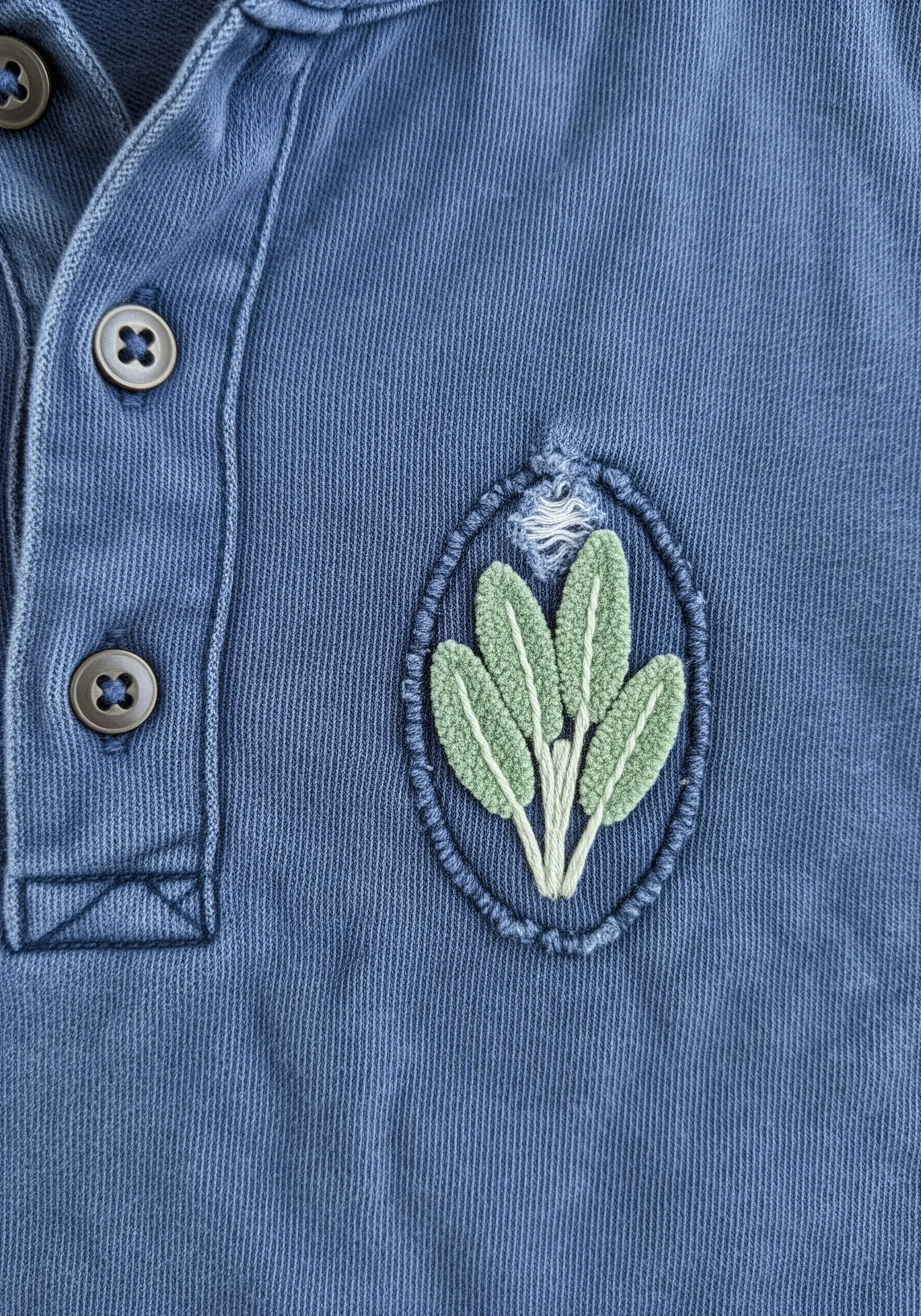 A small oval patch on a blue polo shirt with fuzzy, textured sage leaves embroidered inside.