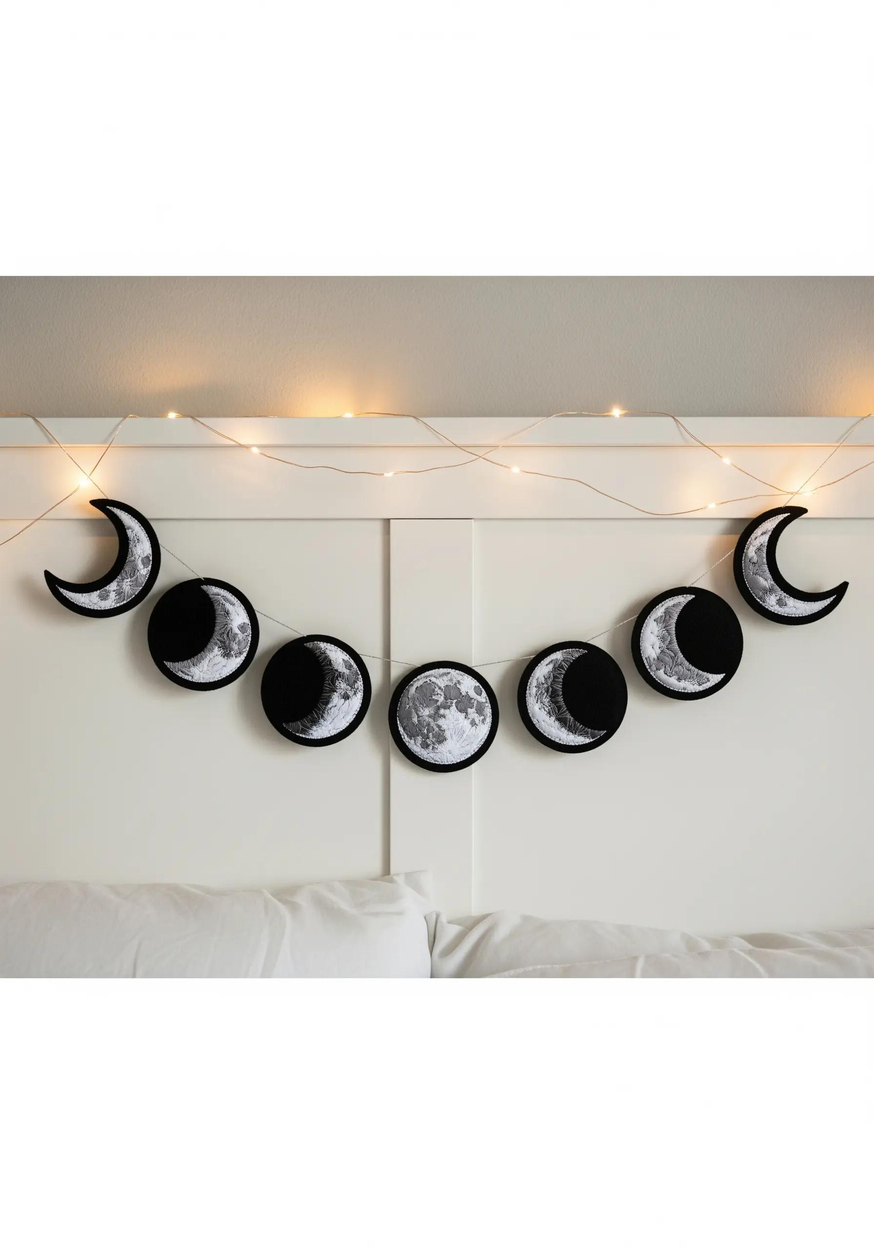 An embroidered moon phase garland hanging above a white headboard.