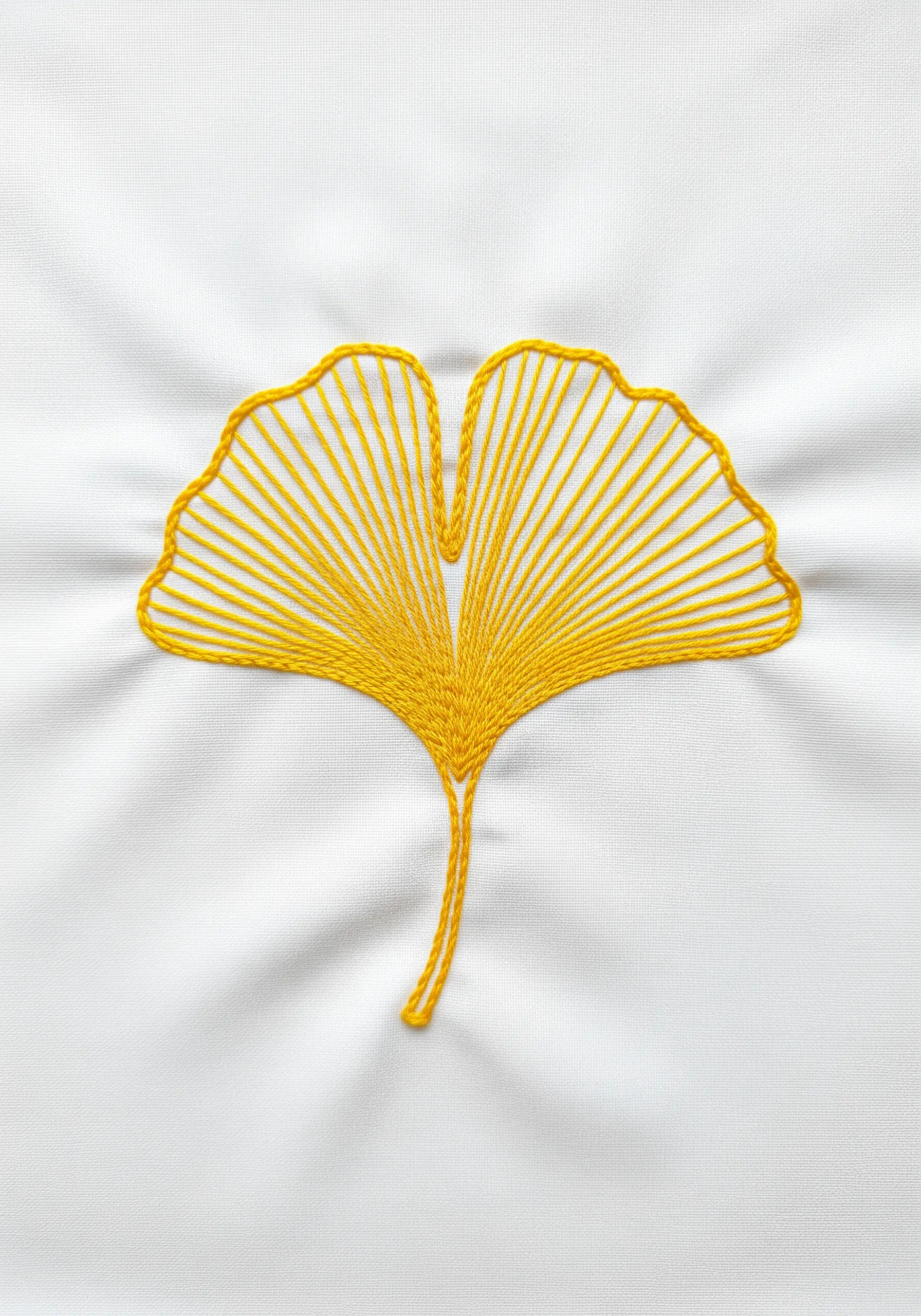 A ginkgo leaf embroidered with radiating yellow straight stitches on white fabric.