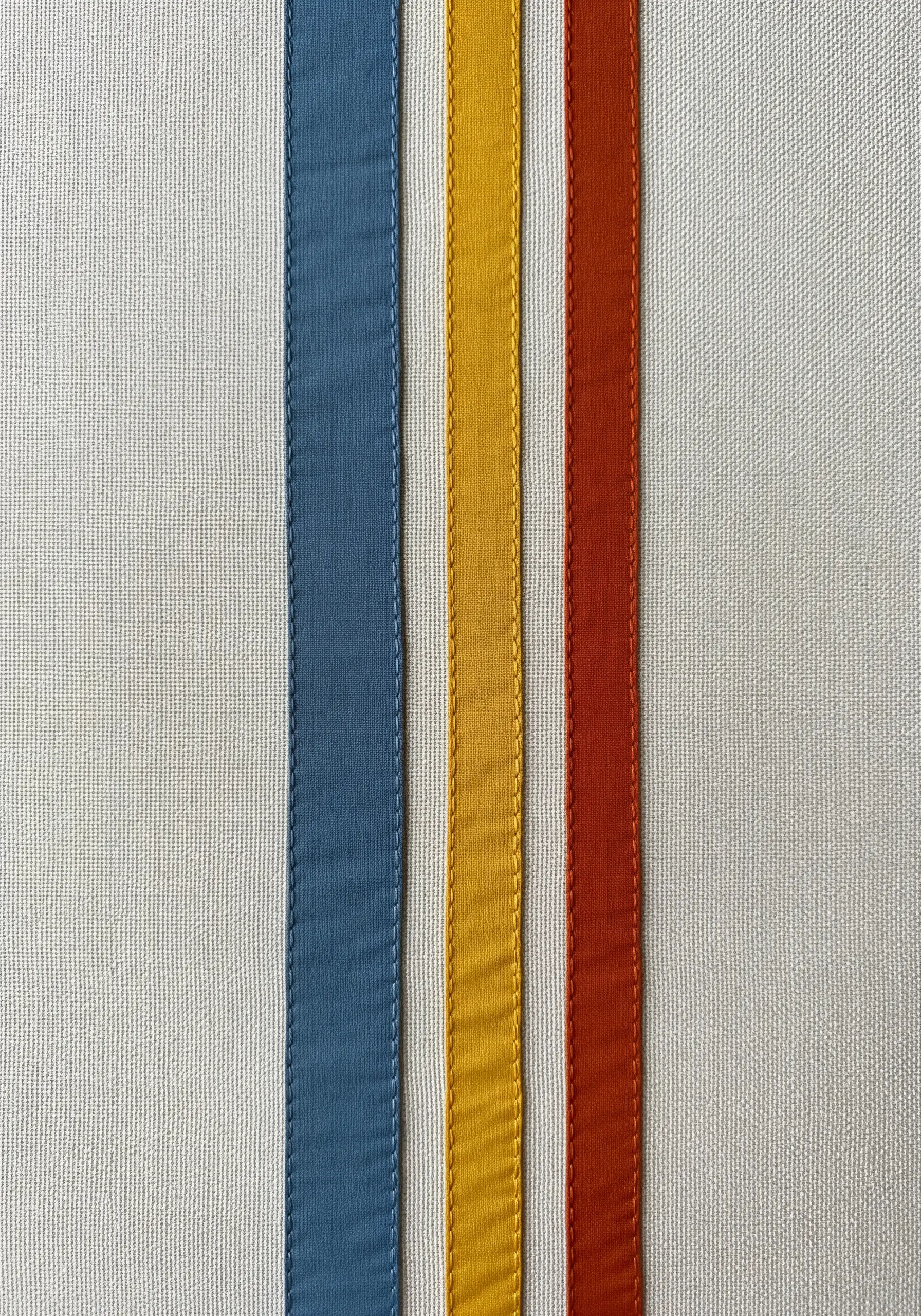 Three vertical stripes of colored fabric—blue, yellow, and rust—stitched onto a white background.