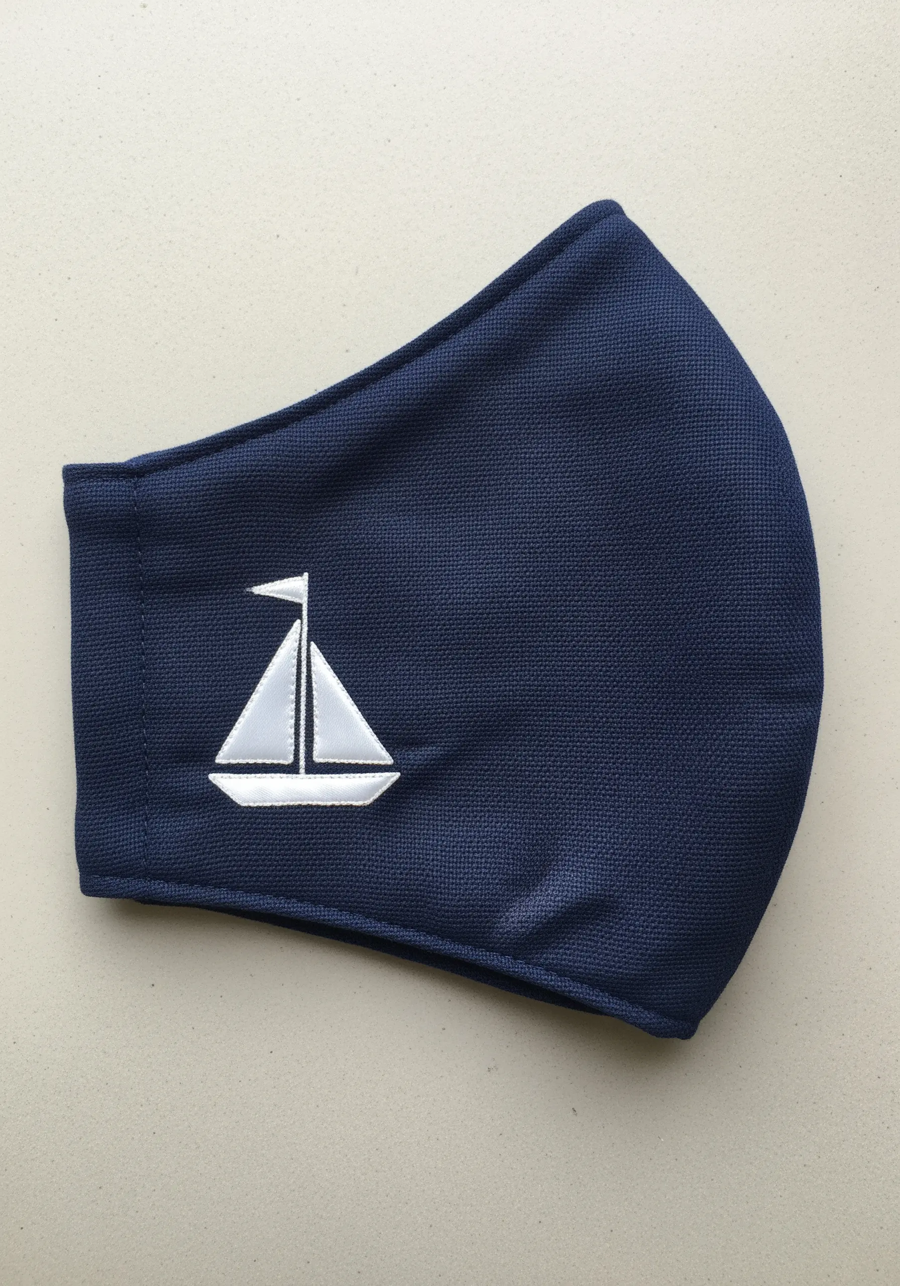 A white sailboat with a grey outline embroidered on a dark blue fabric face mask.