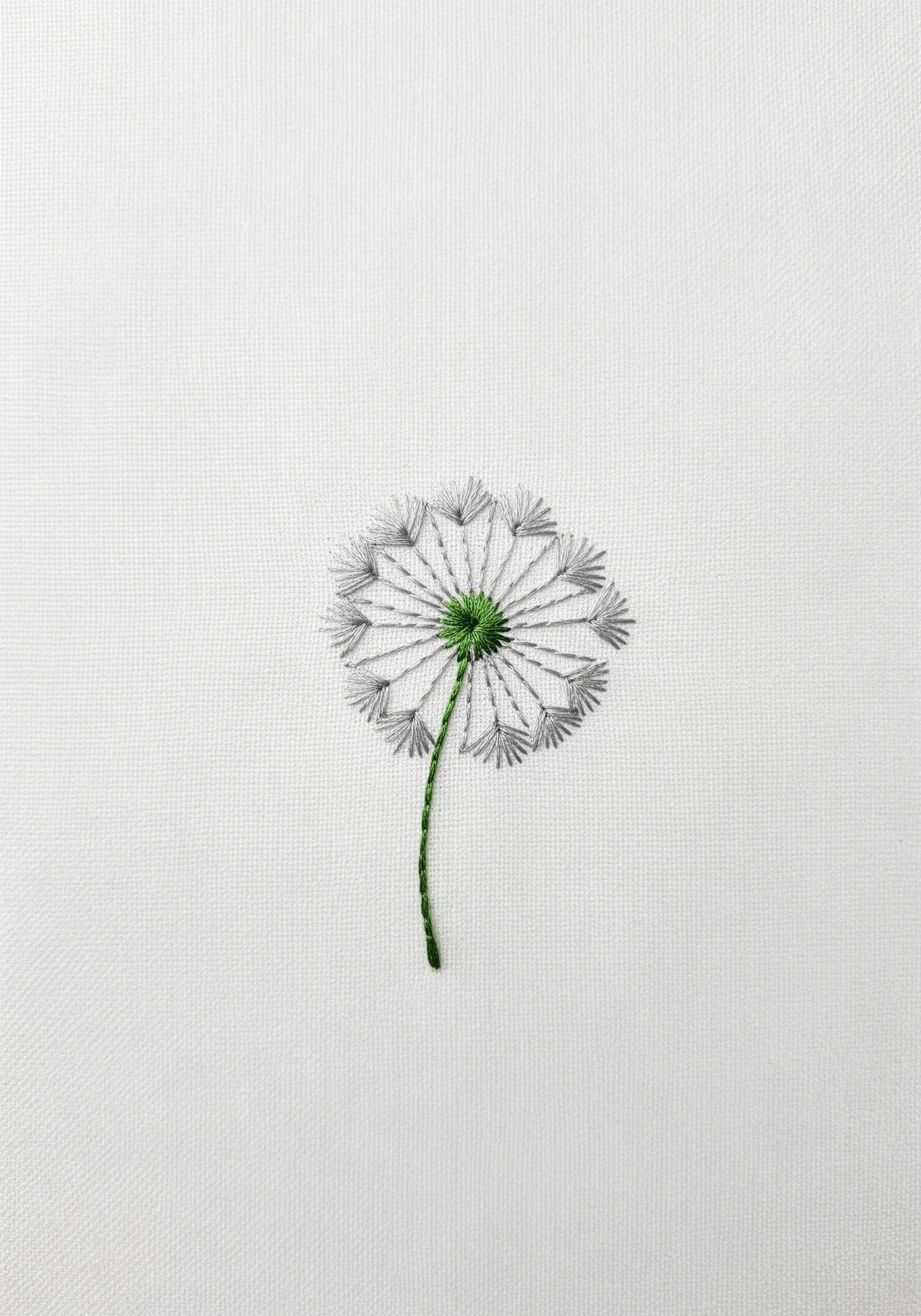 A delicate dandelion seed head stitched with grey and green thread on white fabric.