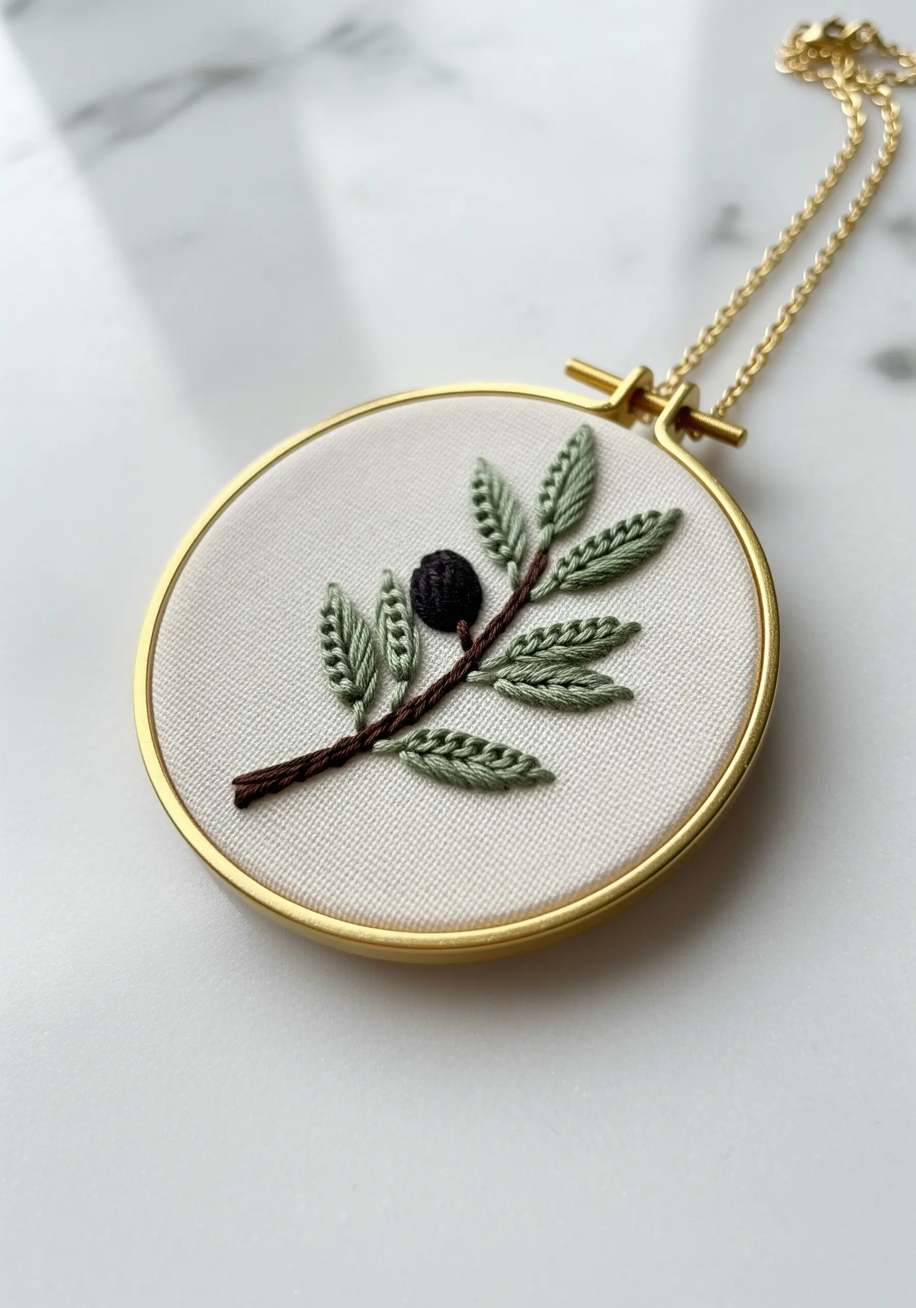 A minimalist embroidered olive branch with varied leaf stitches in a gold pendant.