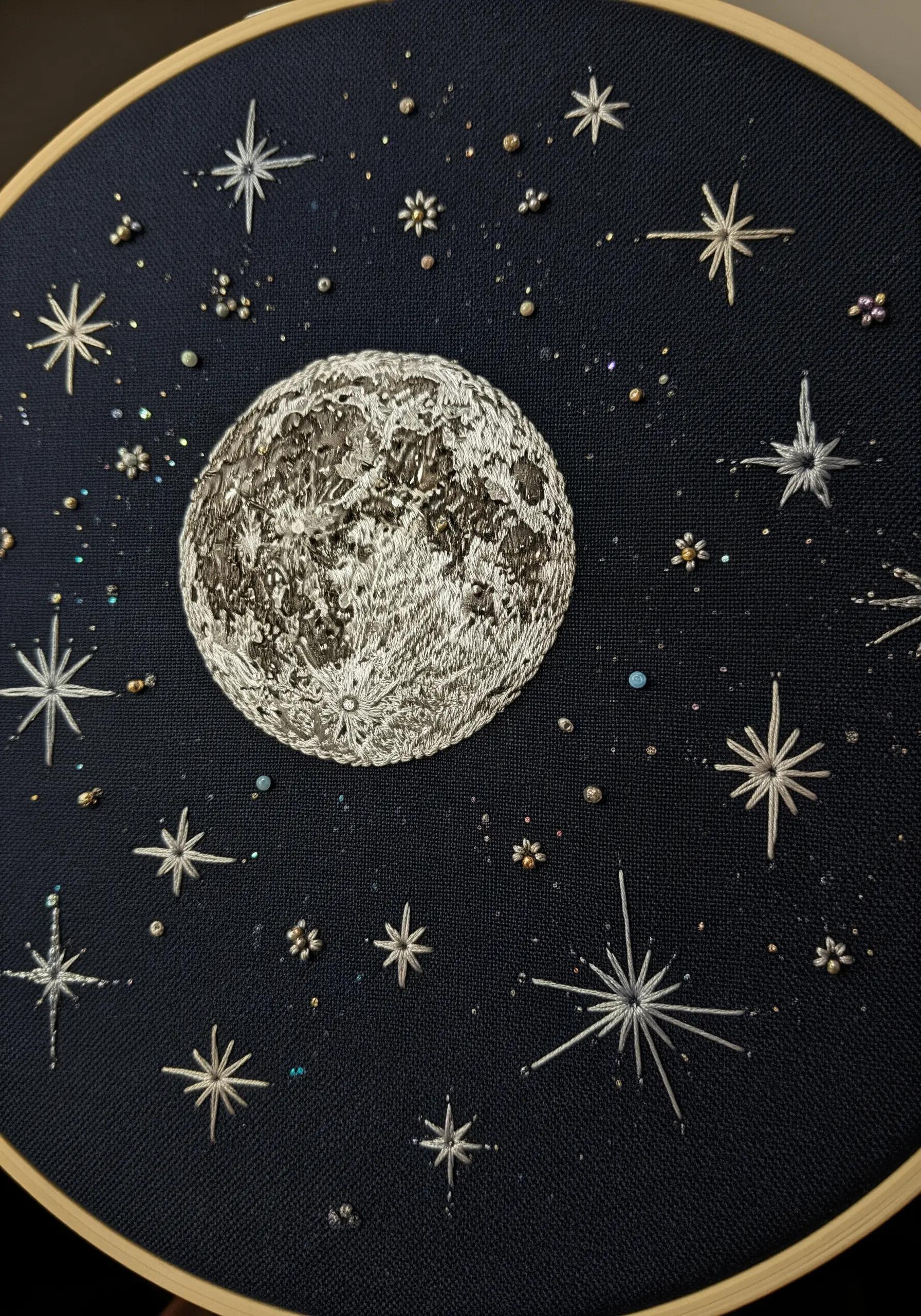 A detailed hand embroidery of the moon and stars on dark navy fabric in a wooden hoop.
