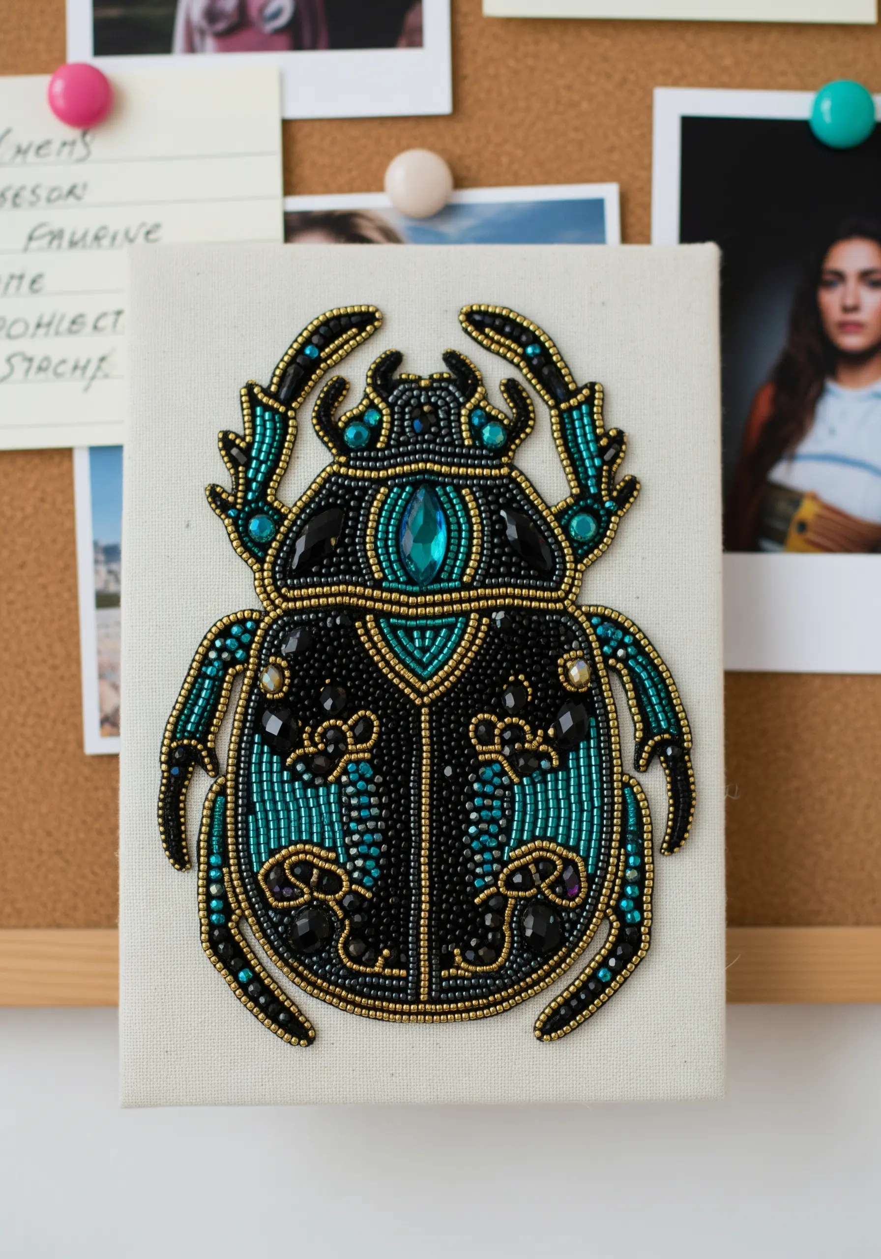 An intricate bead embroidery piece of a scarab beetle in teal, black, and gold.