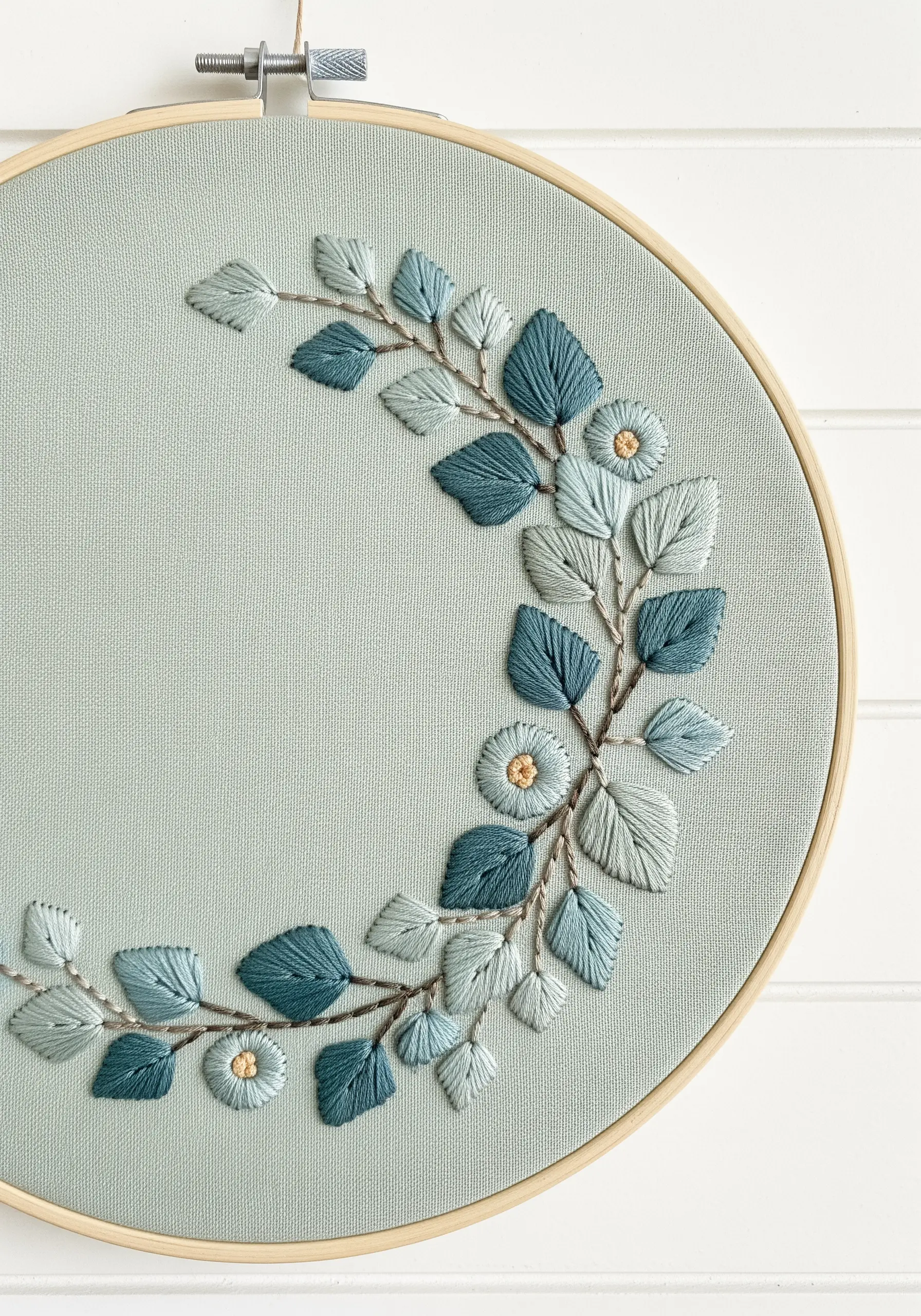 Detail of a floral wreath embroidery with blue and sage satin stitch leaves on muted green fabric.