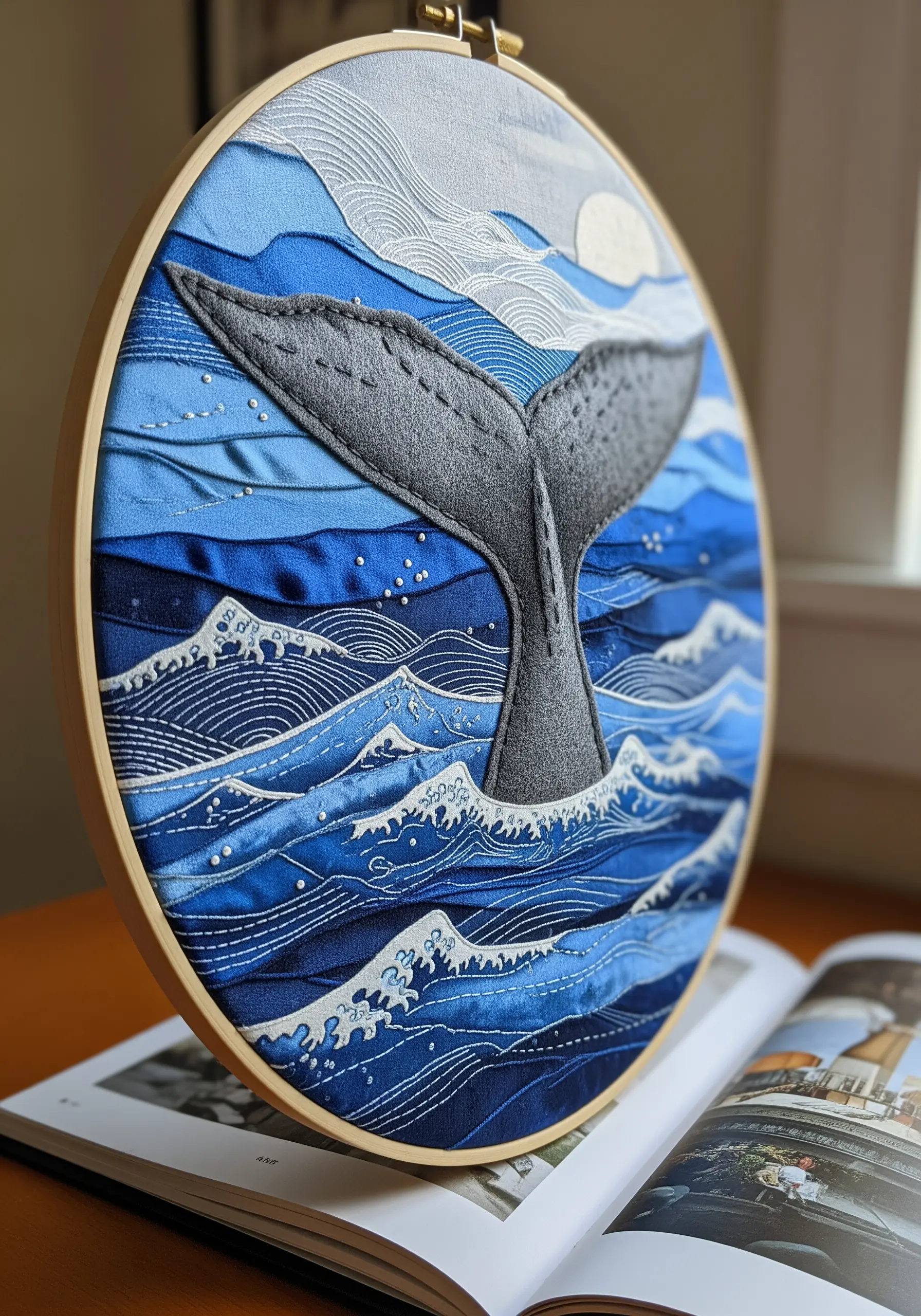 Fabric collage hoop art of a whale tail diving into ruched blue silk and satin waves.