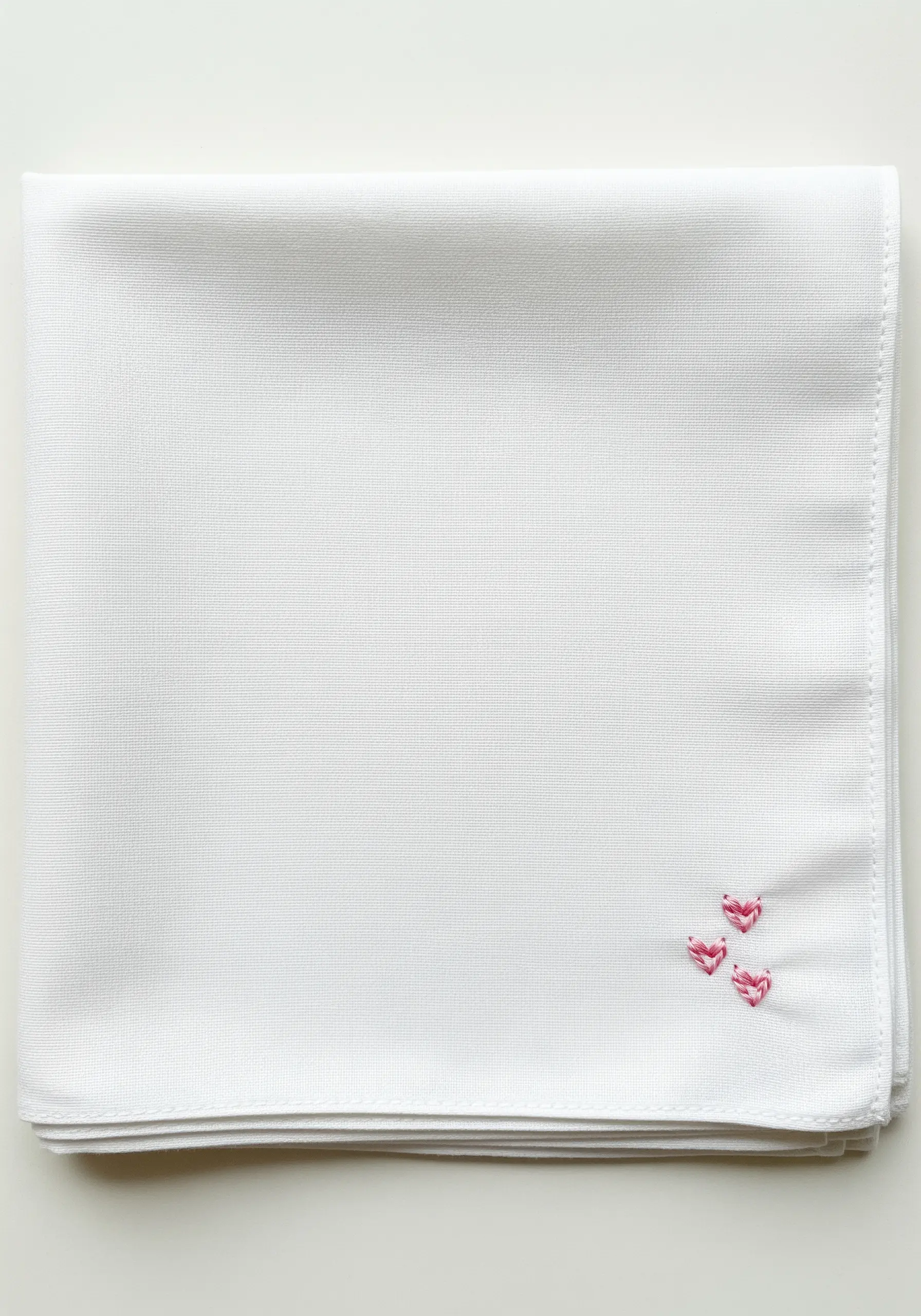 Three tiny, pink, padded satin stitch hearts in the corner of a white handkerchief.