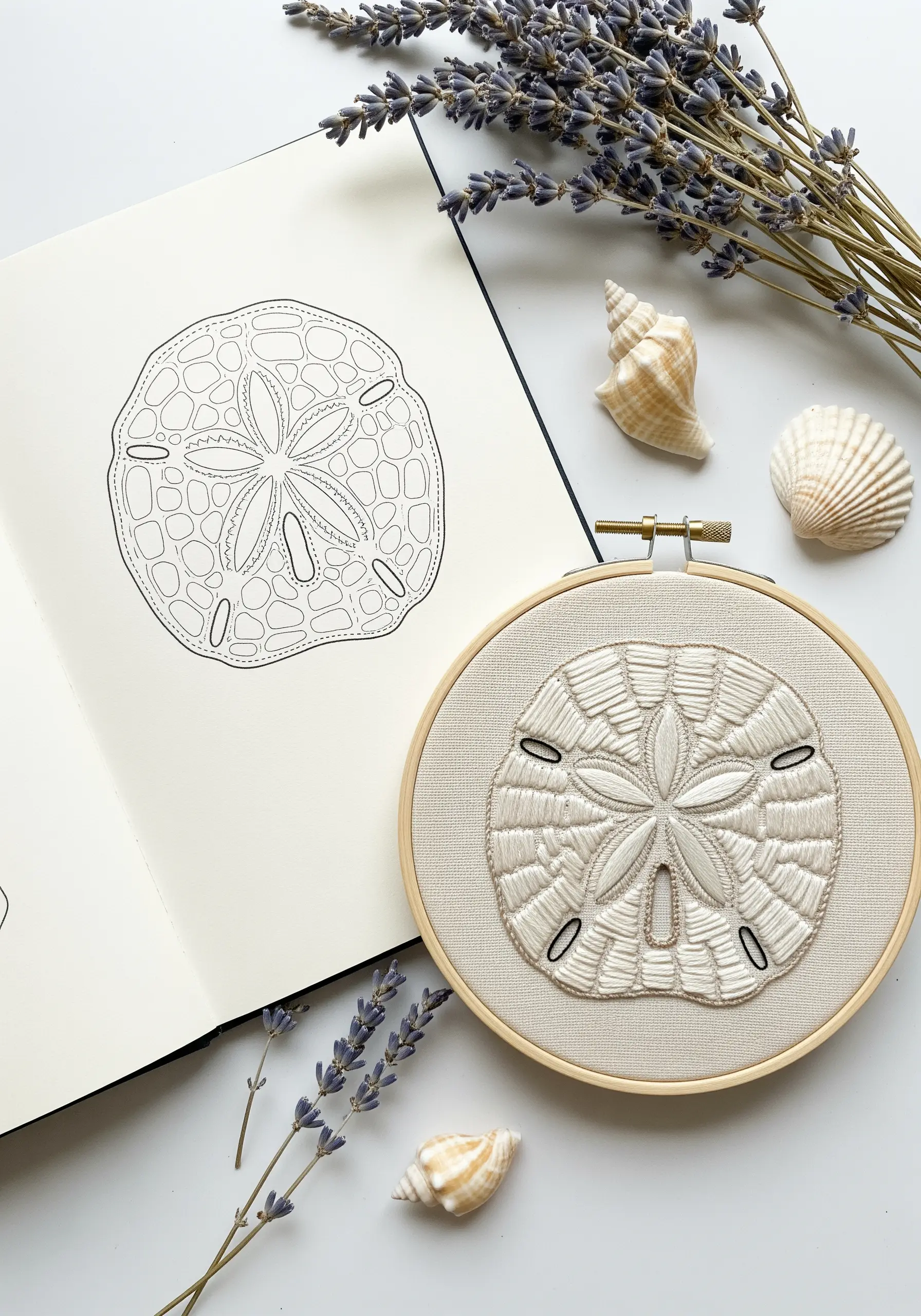 An embroidered sand dollar in a hoop, with the original sketch visible in a notebook.