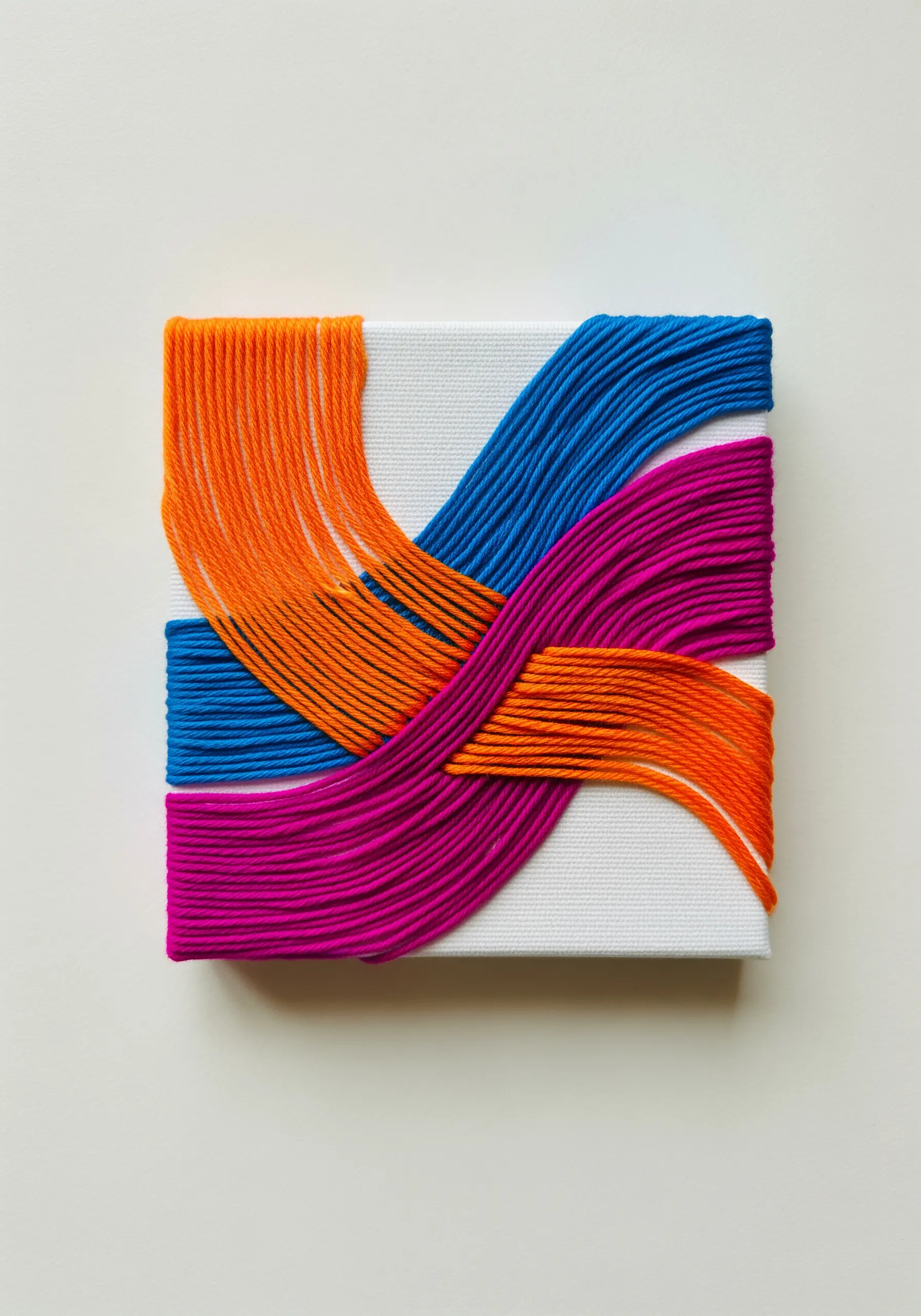 A small canvas wrapped in intersecting bands of orange, blue, and magenta yarn.