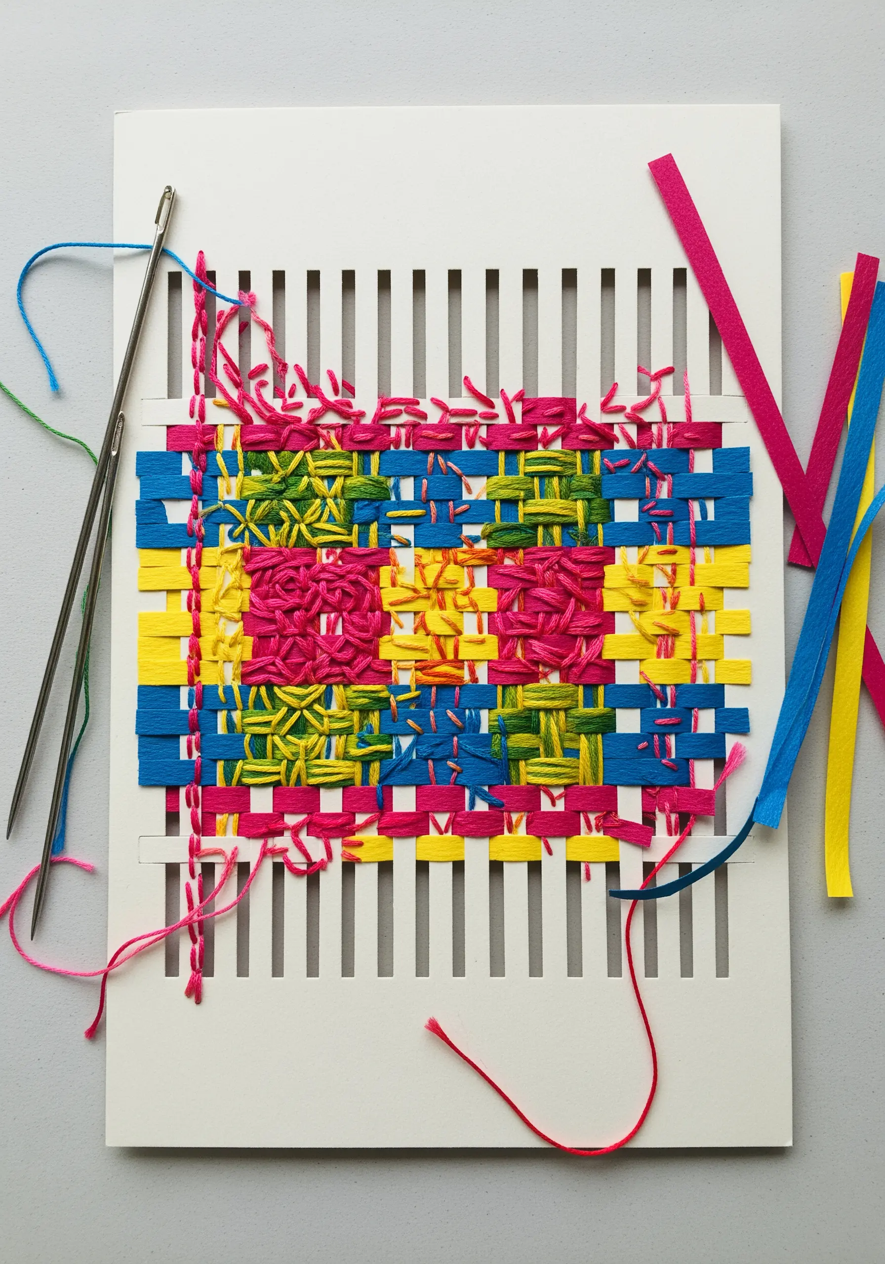 A paper loom woven with blue and pink paper strips, with colorful freestyle embroidery on top.