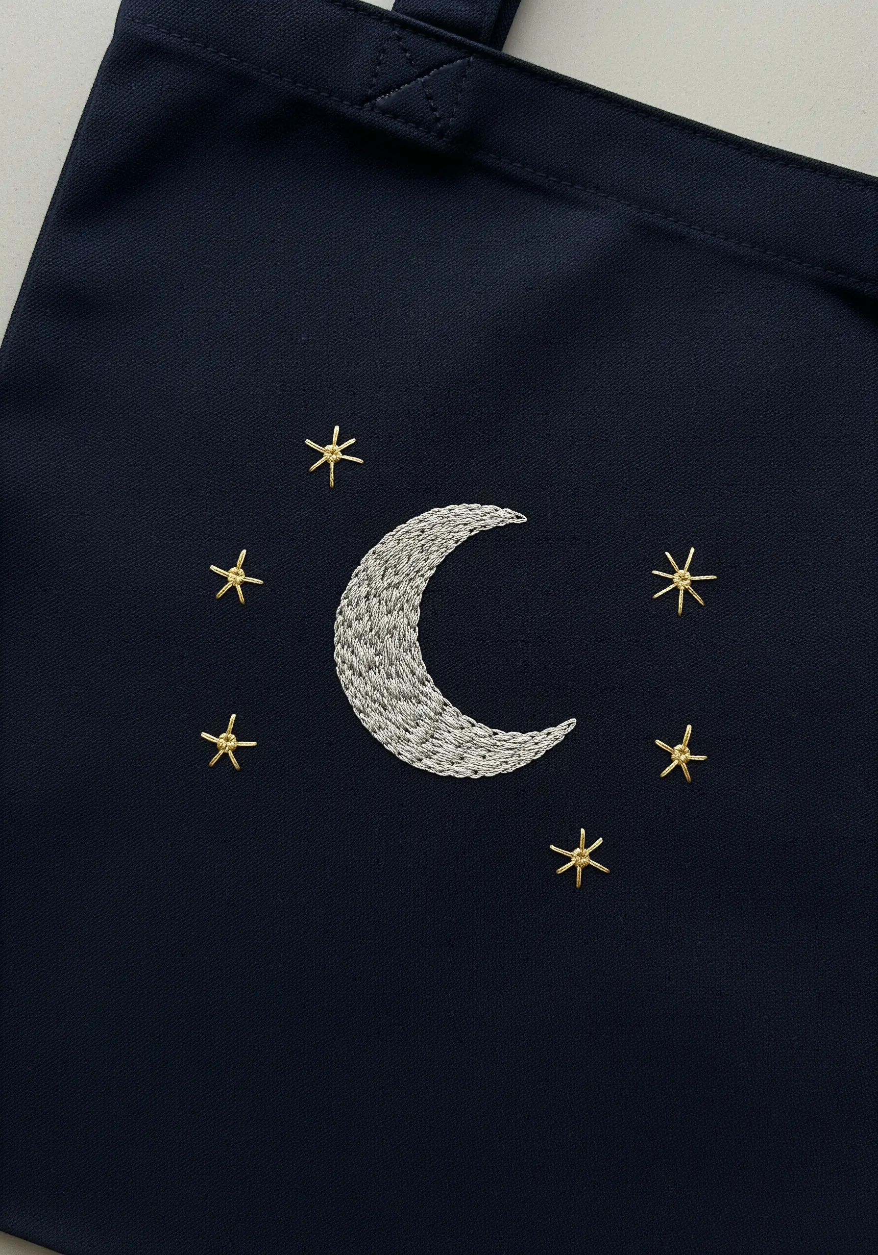 Embroidered crescent moon in silver thread with gold stars on a dark navy tote bag.