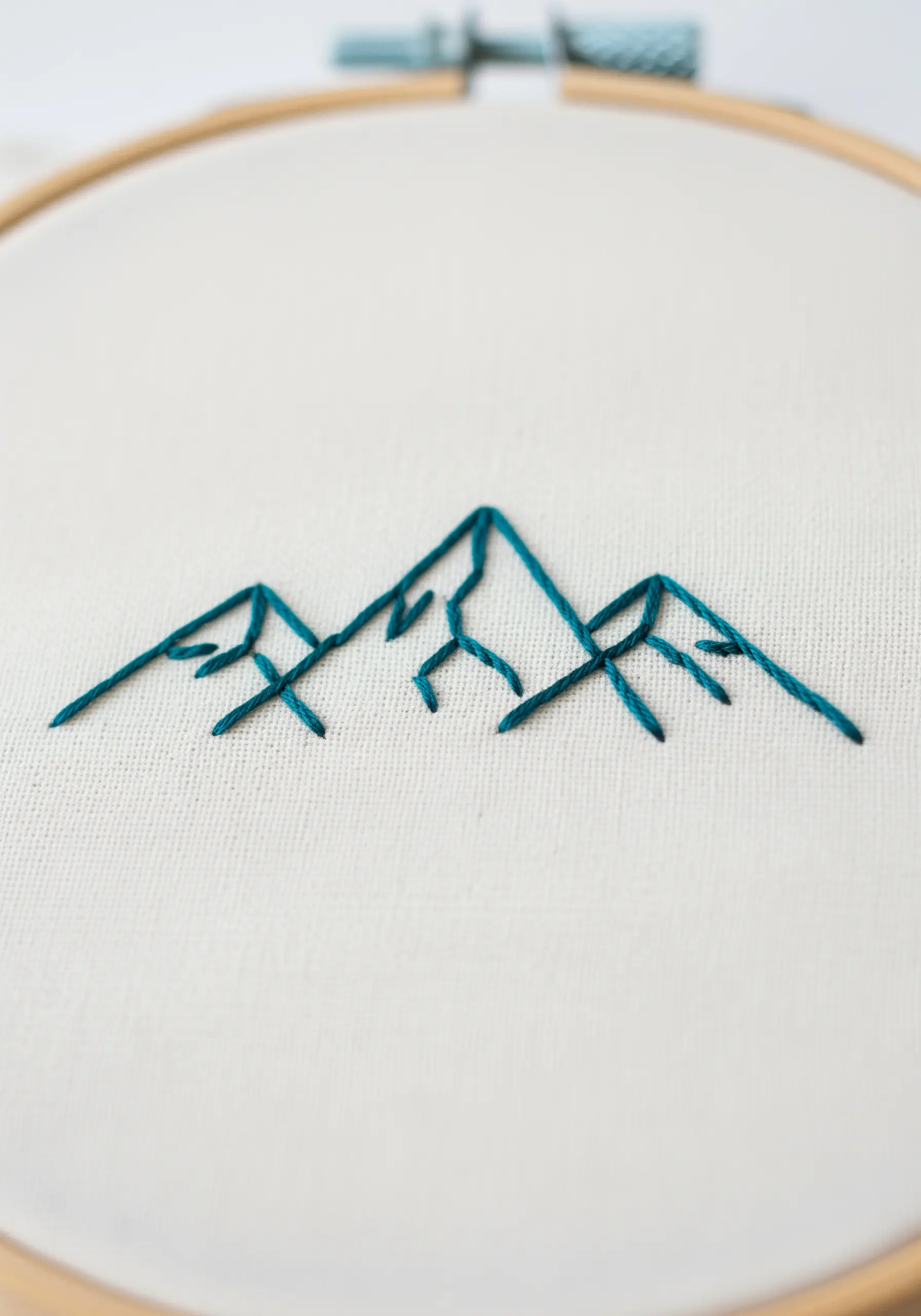 Minimalist teal line art embroidery of a mountain range on white fabric.