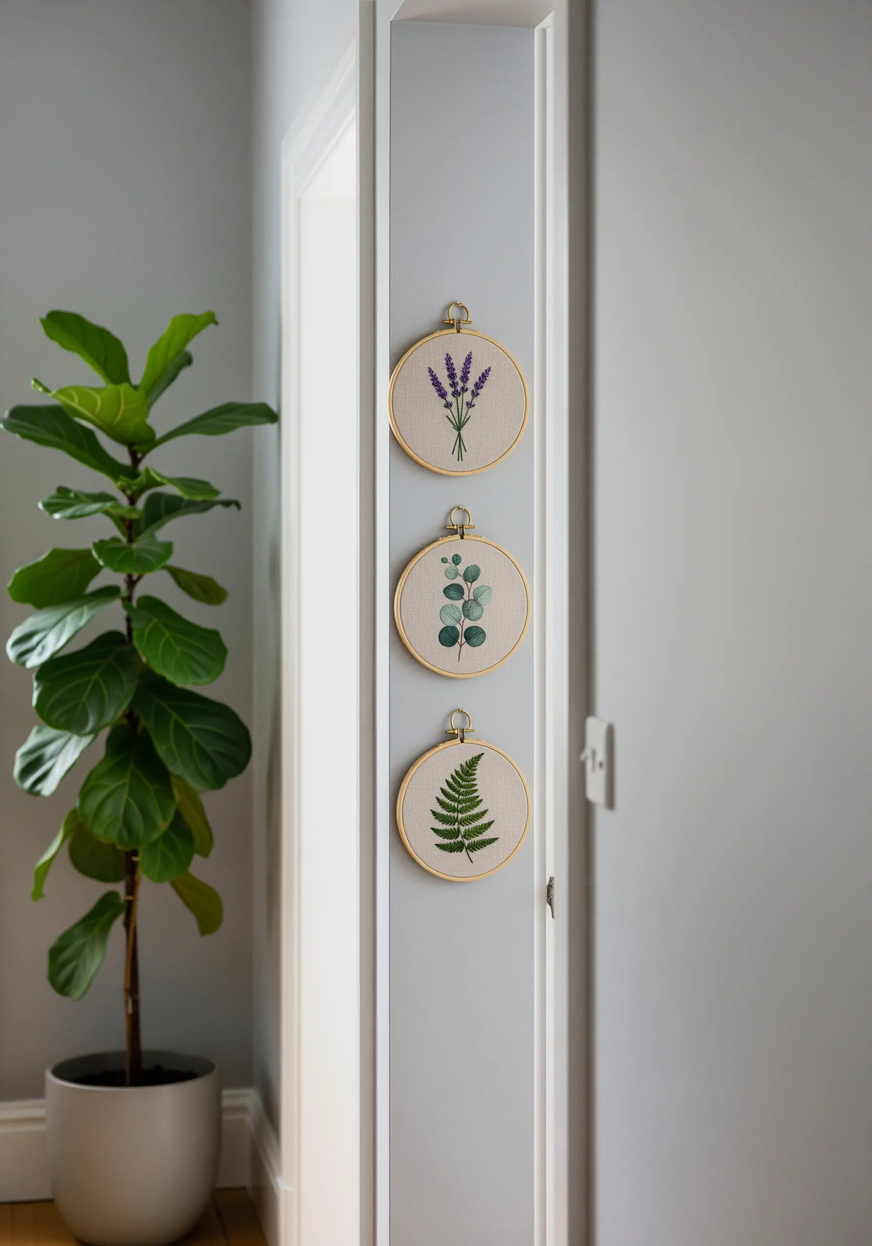 Three embroidery hoops with lavender, eucalyptus, and fern motifs hanging vertically on a gray wall.