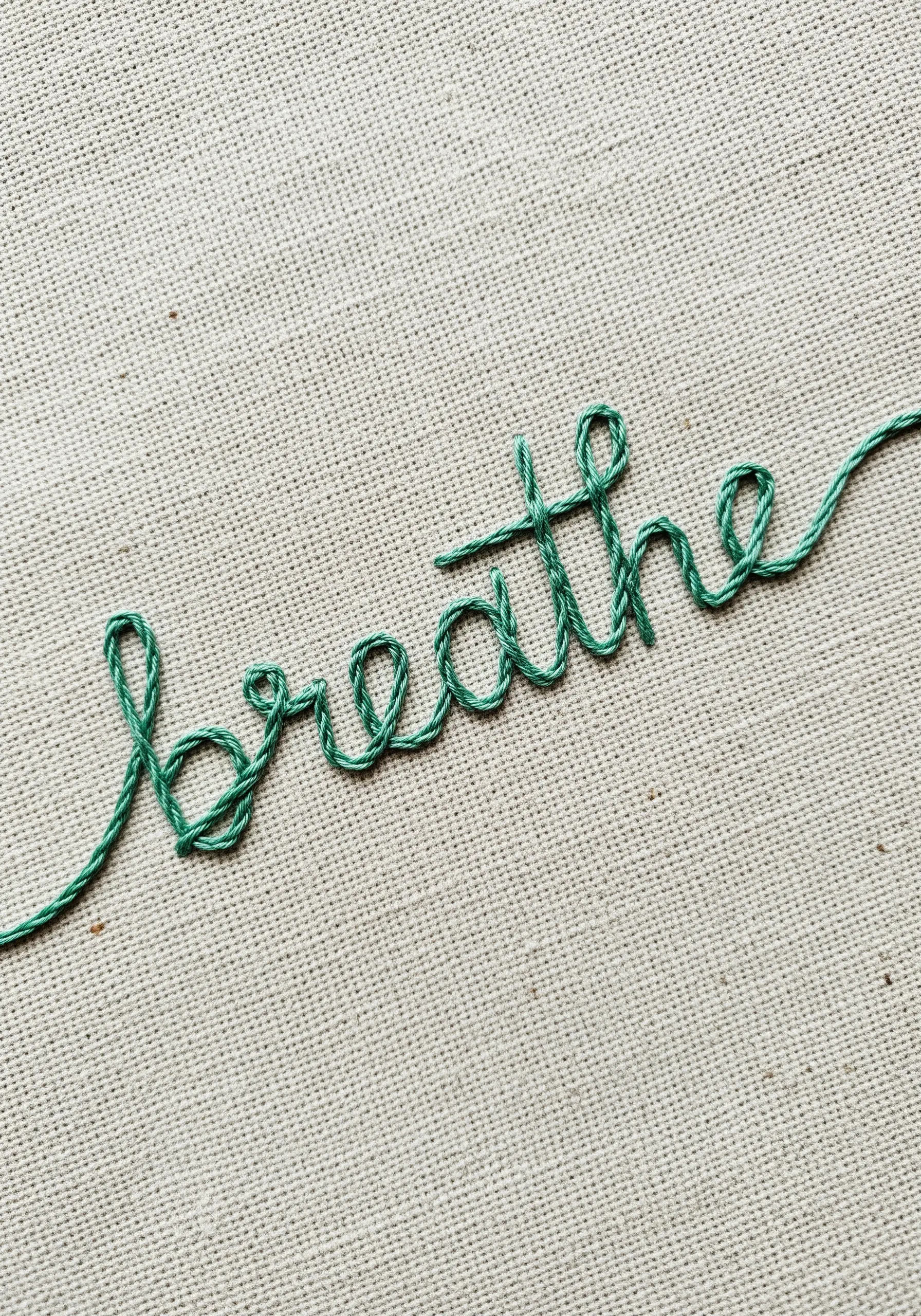 The word 'breathe' embroidered in a flowing green cursive script on linen fabric.