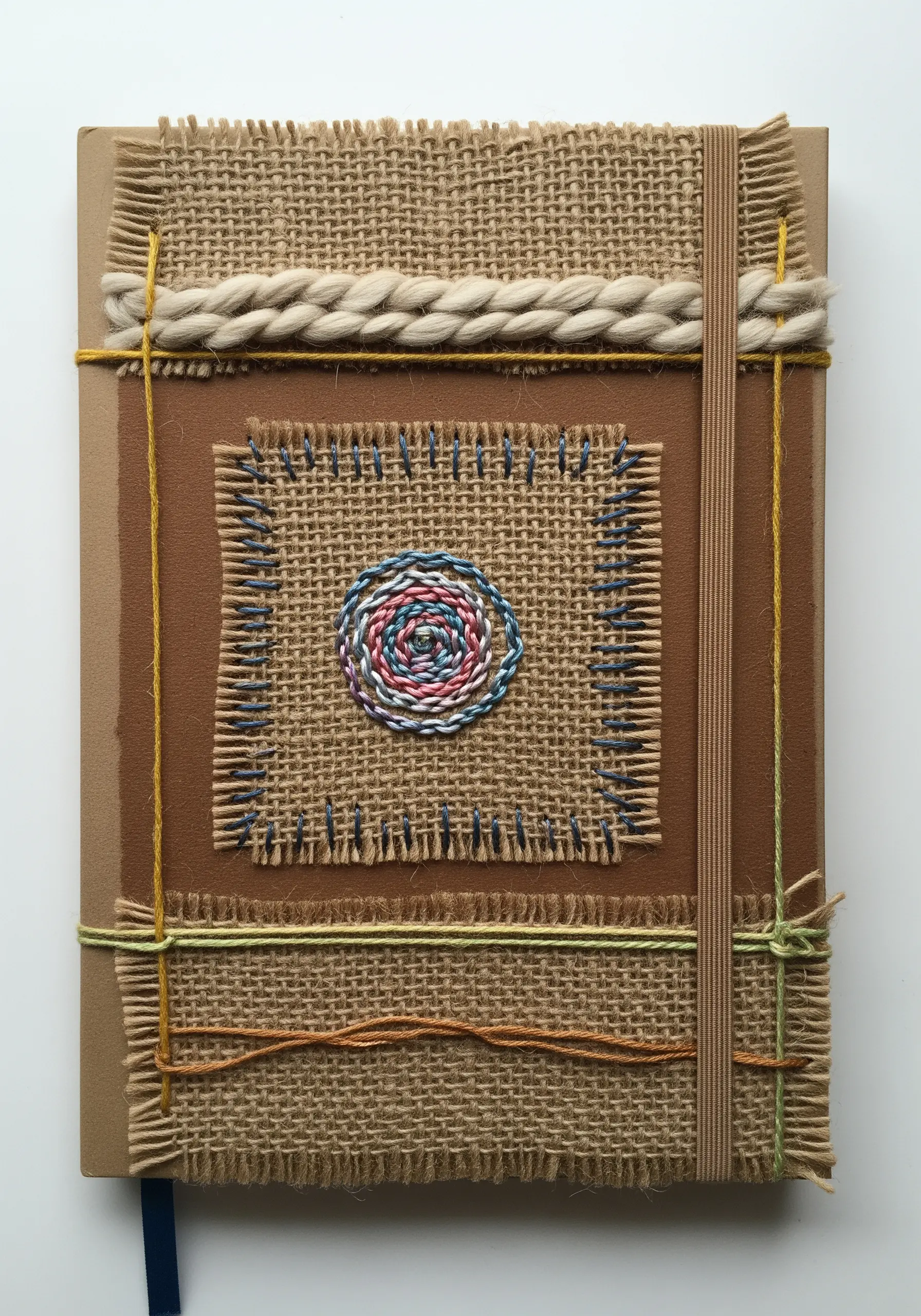A journal cover decorated with layered burlap patches, visible stitching, and a central couched thread spiral.