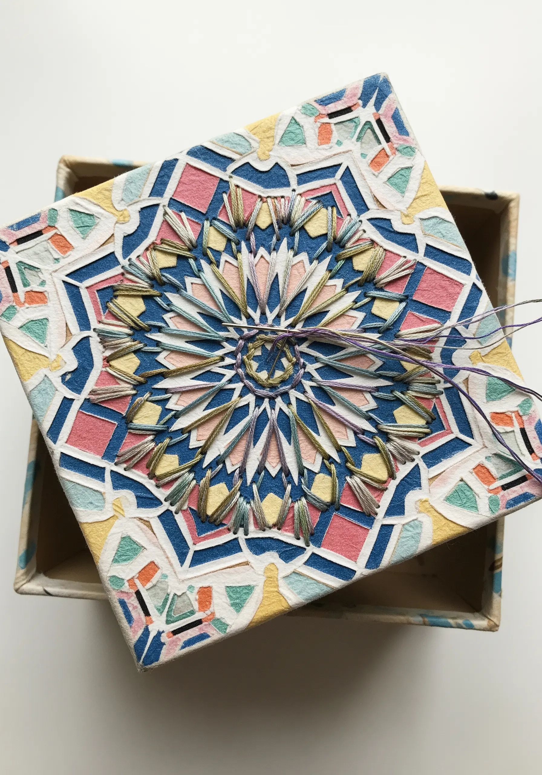 A box lid decorated with a colorful geometric mandala pattern made with stitched thread.