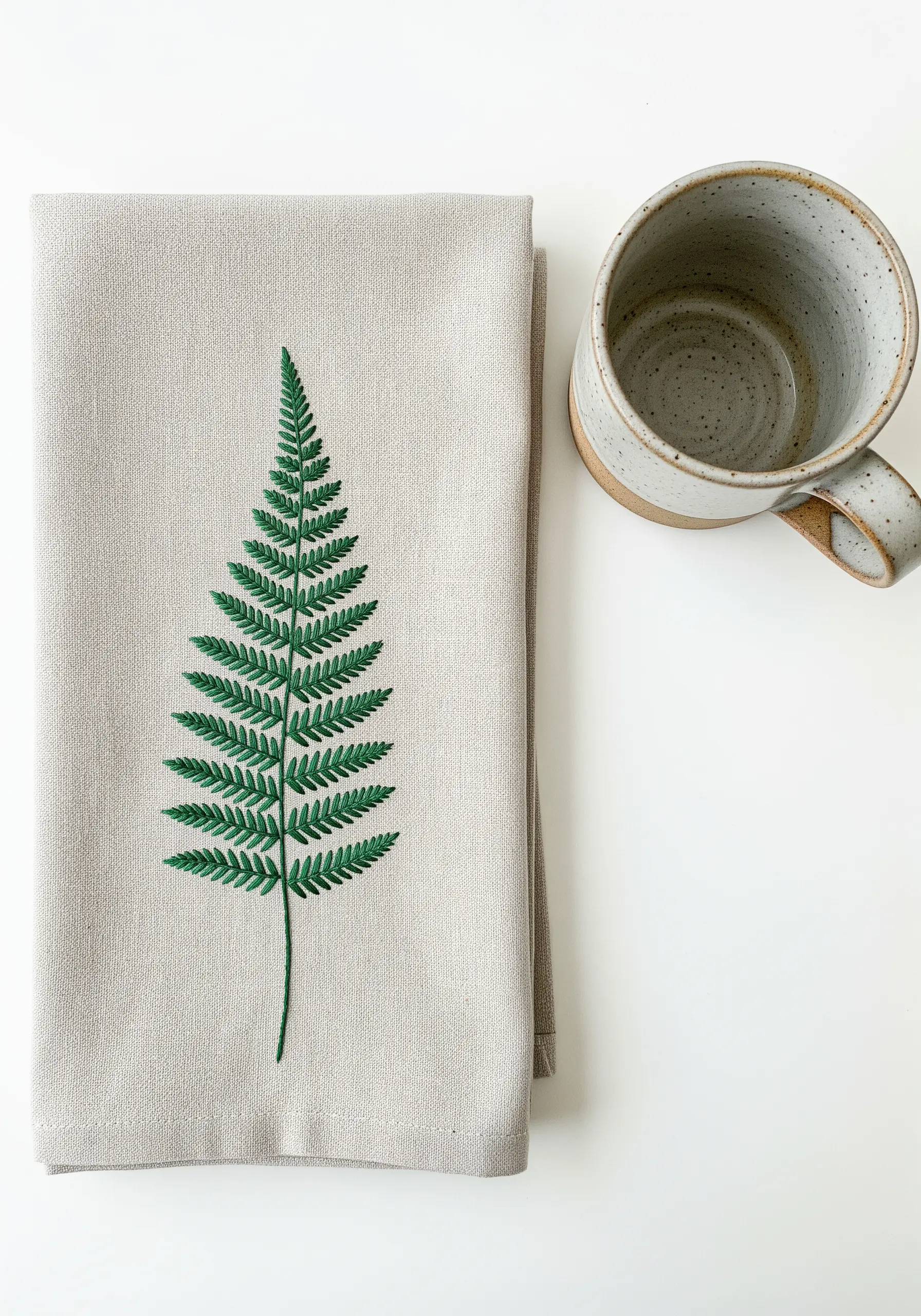 A single, elegant fern frond embroidered in green on a neutral linen tea towel.