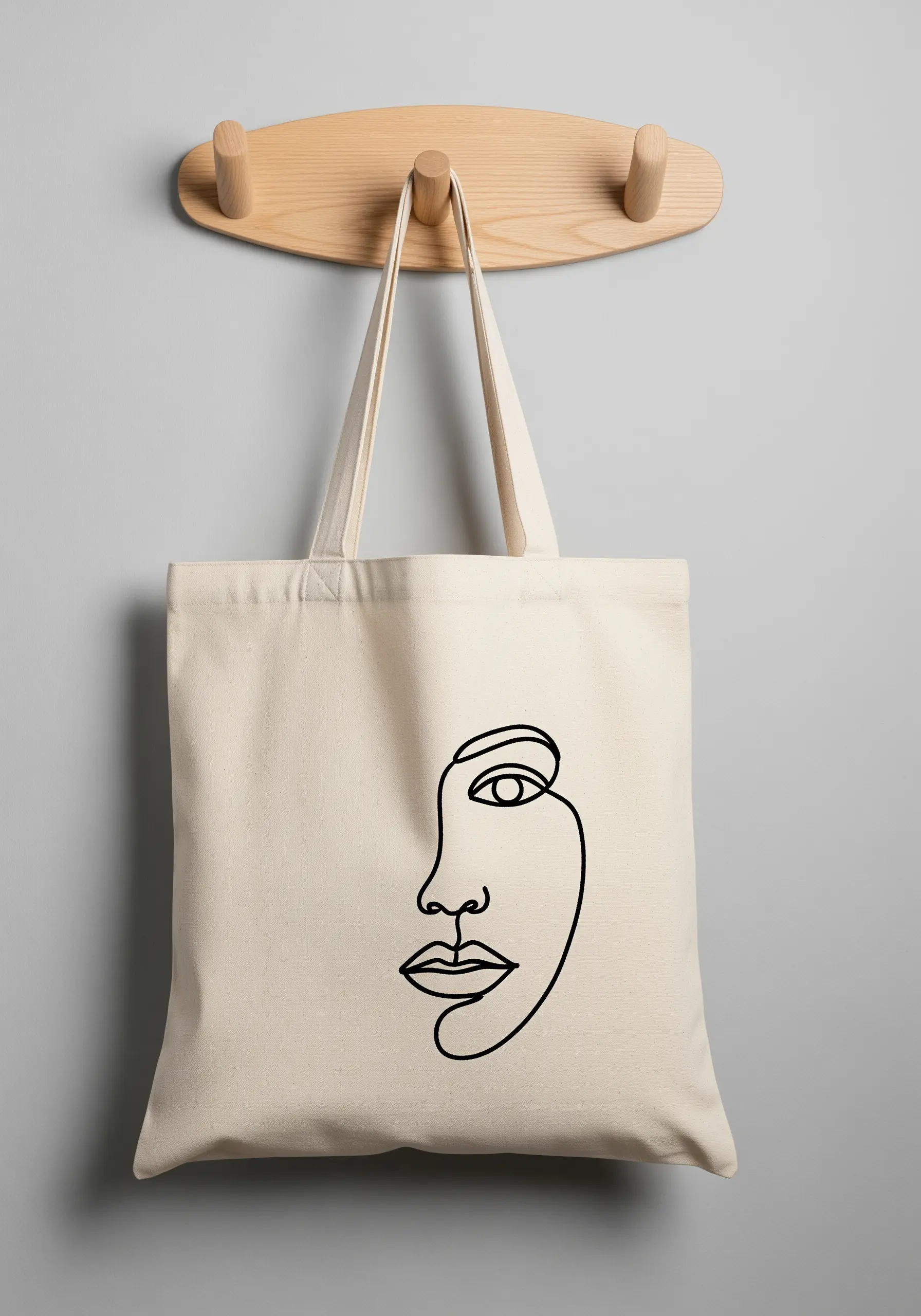 A natural canvas tote bag featuring an abstract face embroidered in a single continuous black line.