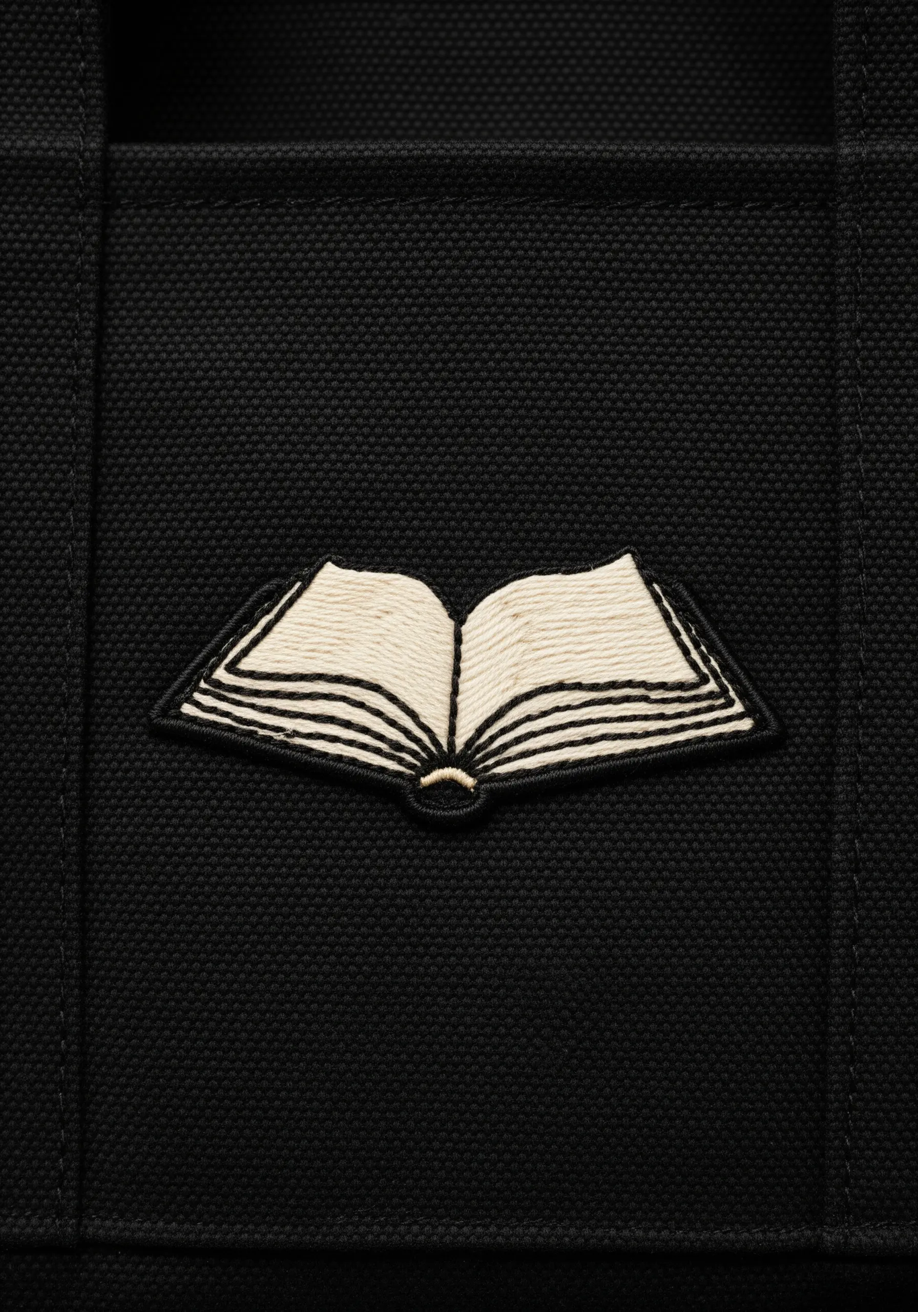 An embroidered open book with a bold black outline and fine lines for pages on a black tote.