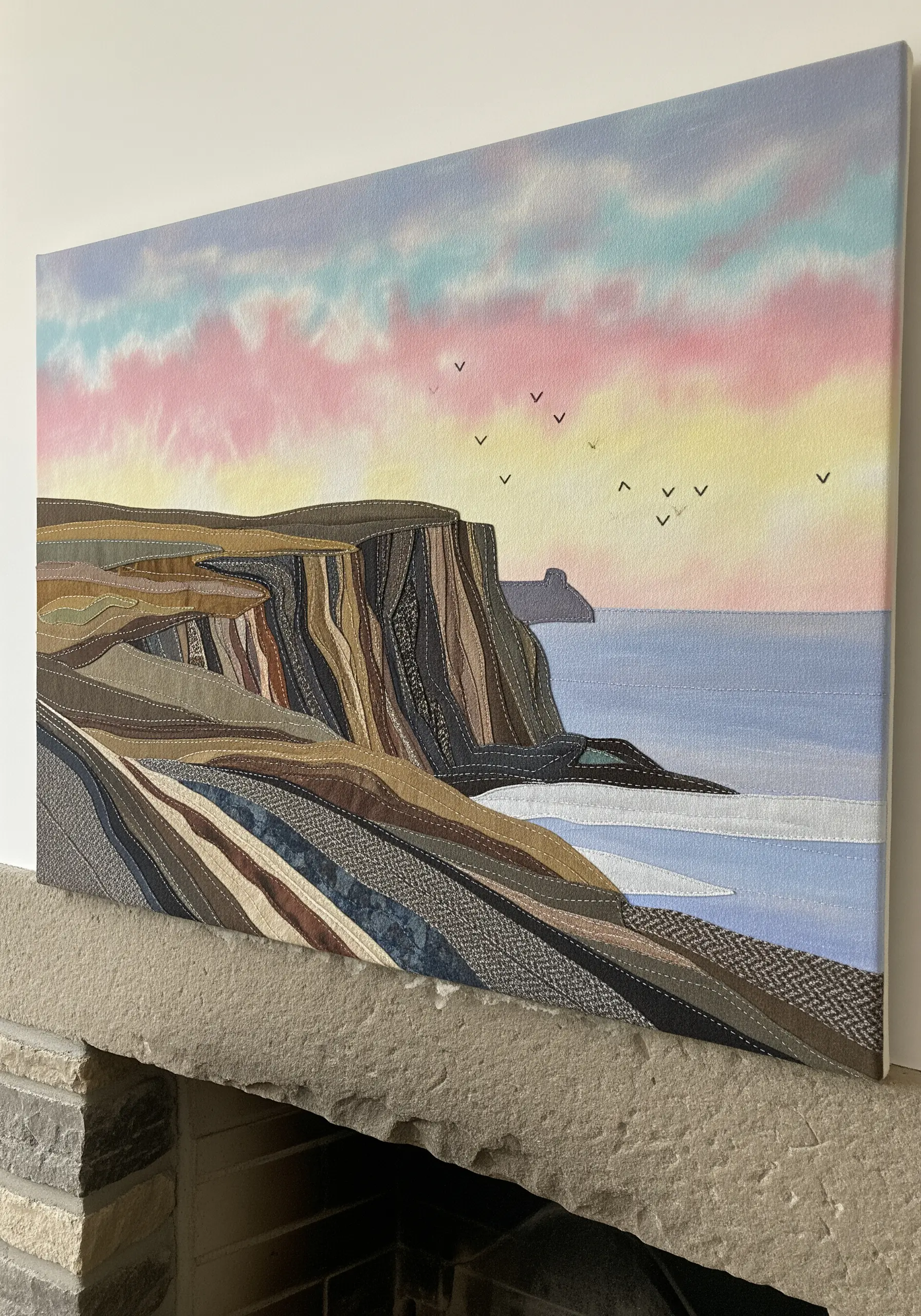 A fabric collage of coastal cliffs at sunset, with visible running stitches defining the rock strata.