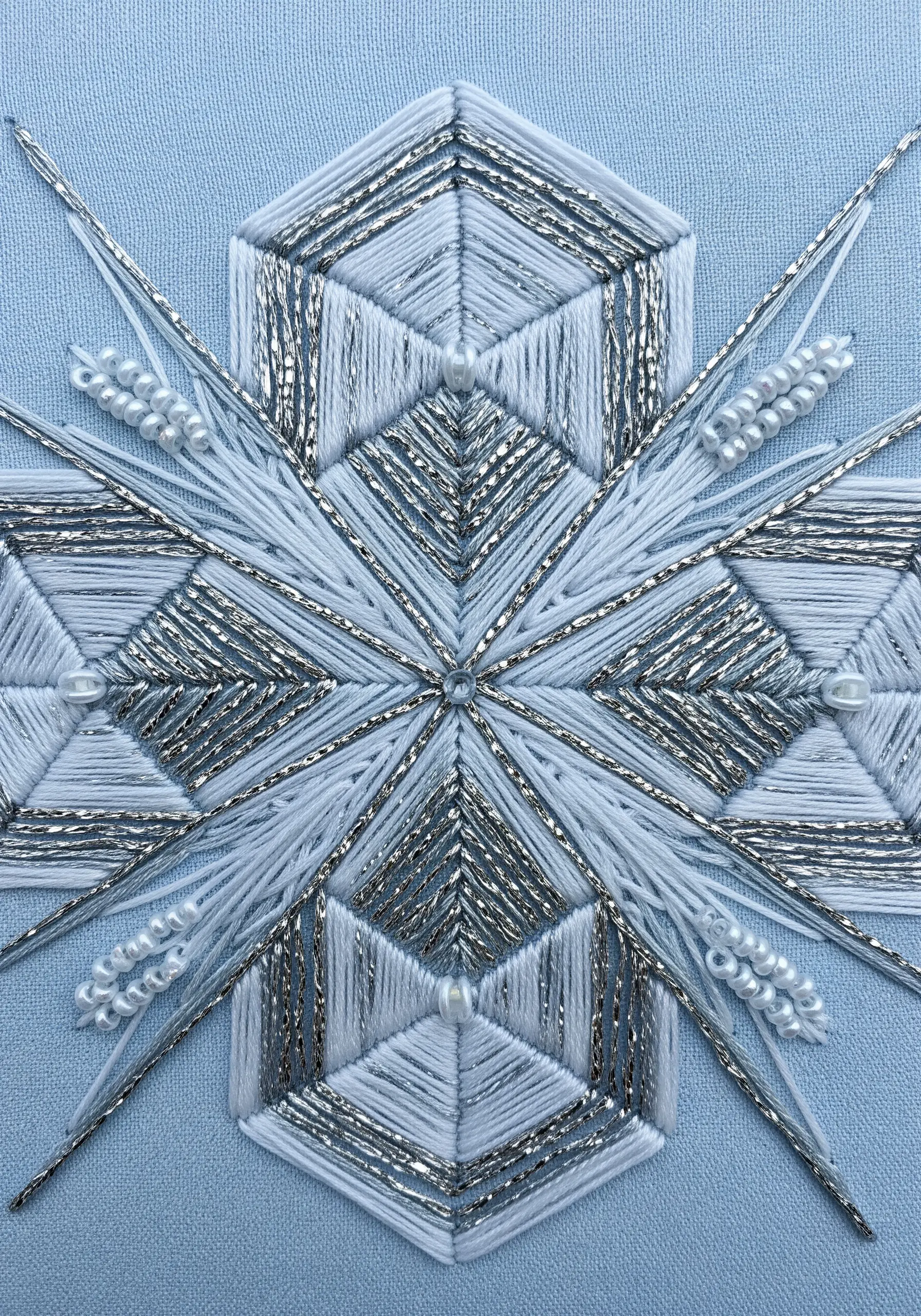 A geometric snowflake embroidered with white and silver metallic thread, accented with small pearls.