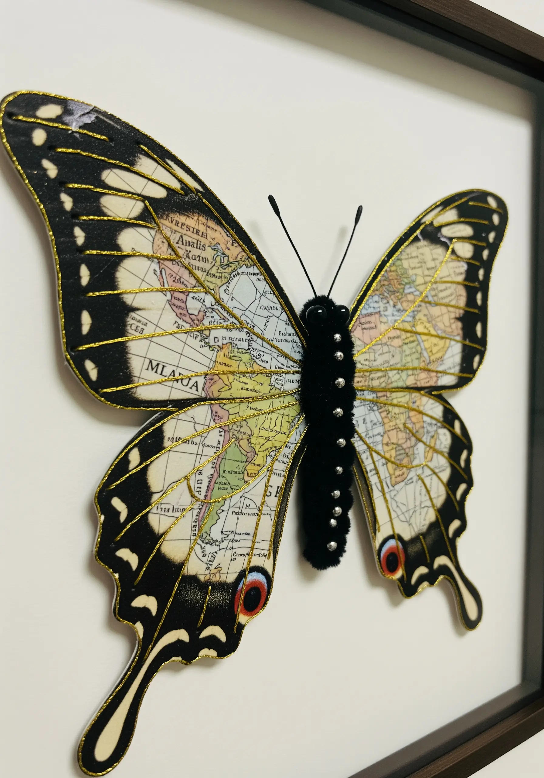 A framed paper butterfly with a map pattern on its wings and a textured, embroidered body.