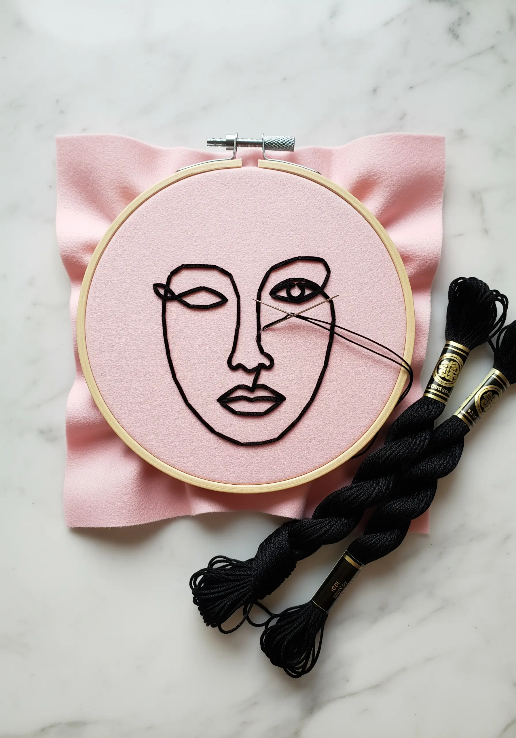 Abstract face outlined in bold black thread on a soft pink background.