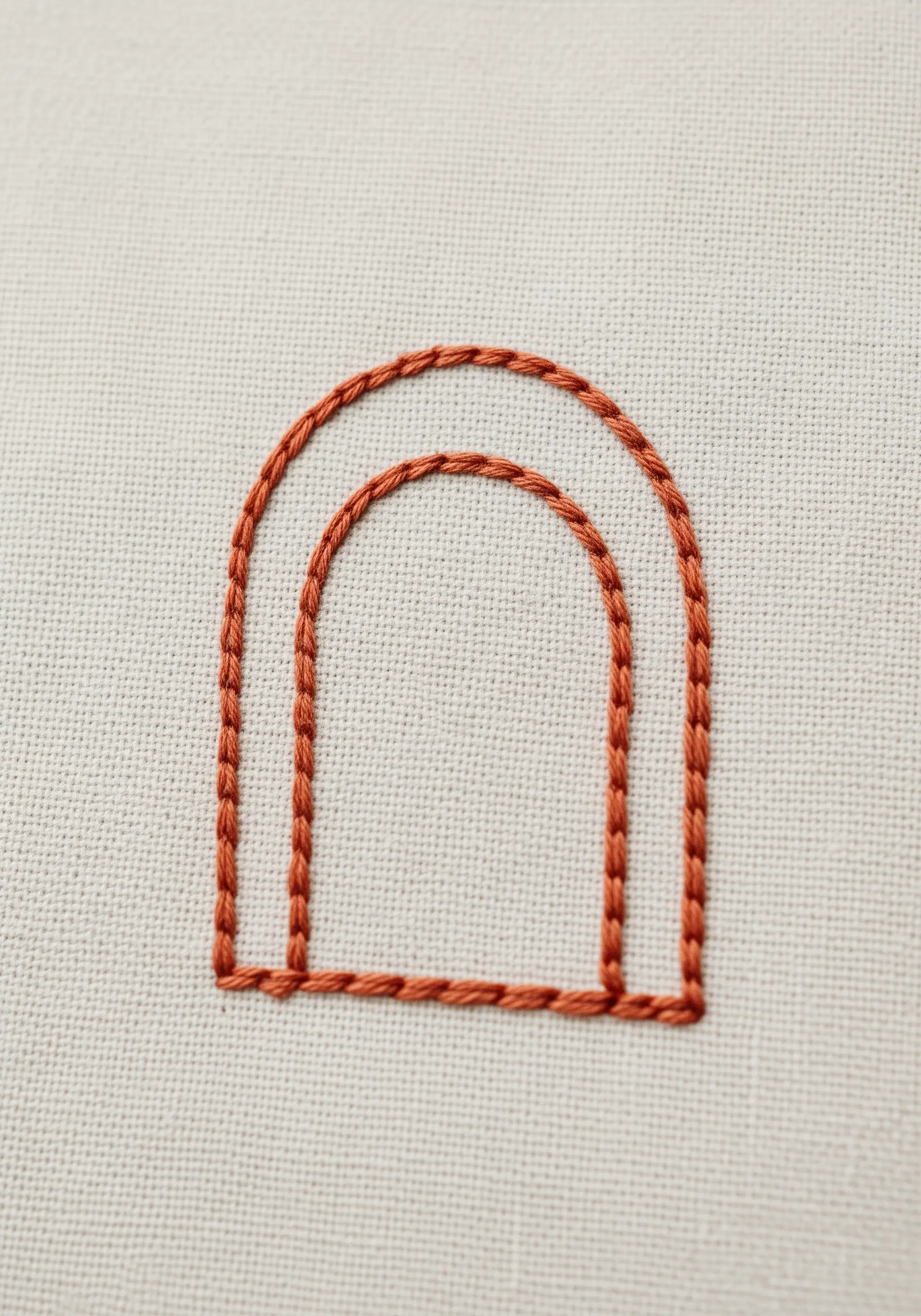 A close-up of a double arch outlined with precise brown split stitch on cream fabric.