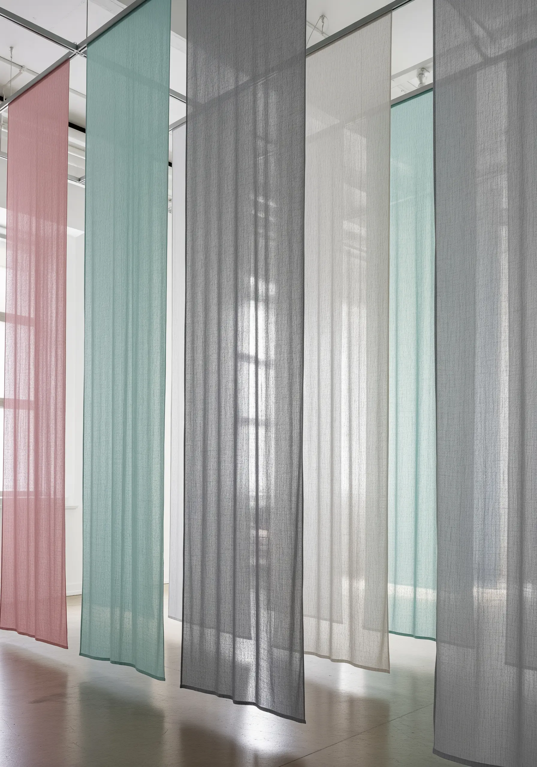 Sheer, floor-length fabric panels in muted tones of pink, seafoam, and gray hanging in a gallery.