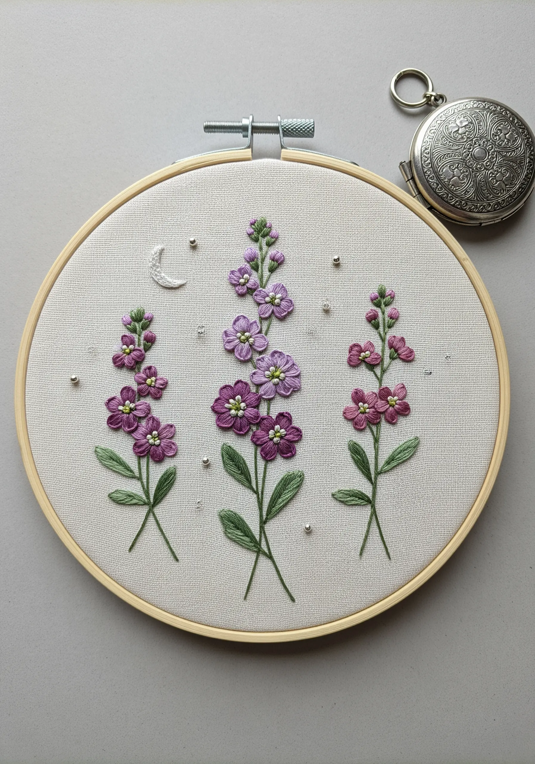 Embroidery of purple and pink night-scented stock with small silver beads on light-colored fabric.