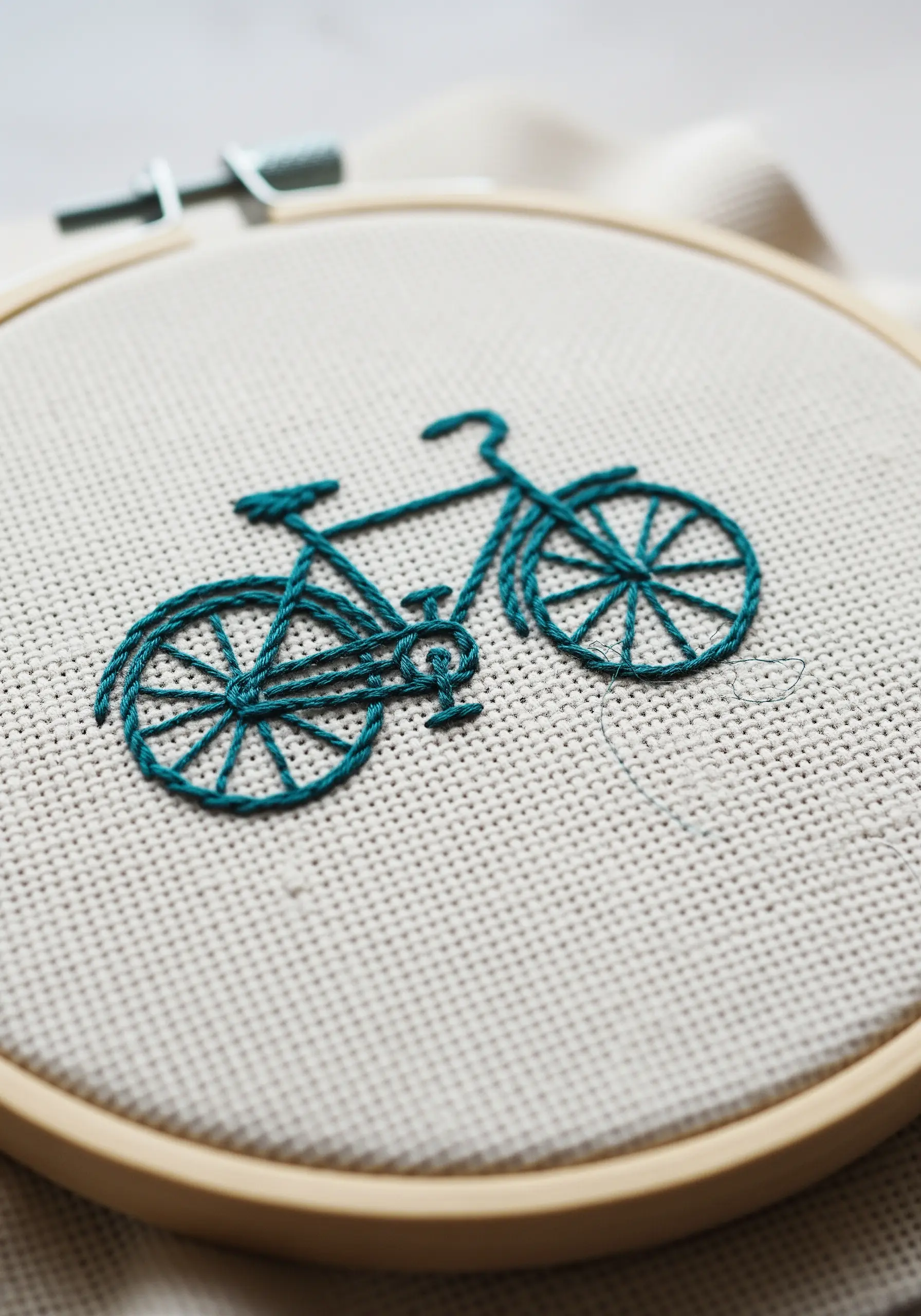 A detailed outline of a bicycle embroidered in teal thread on neutral-colored fabric.