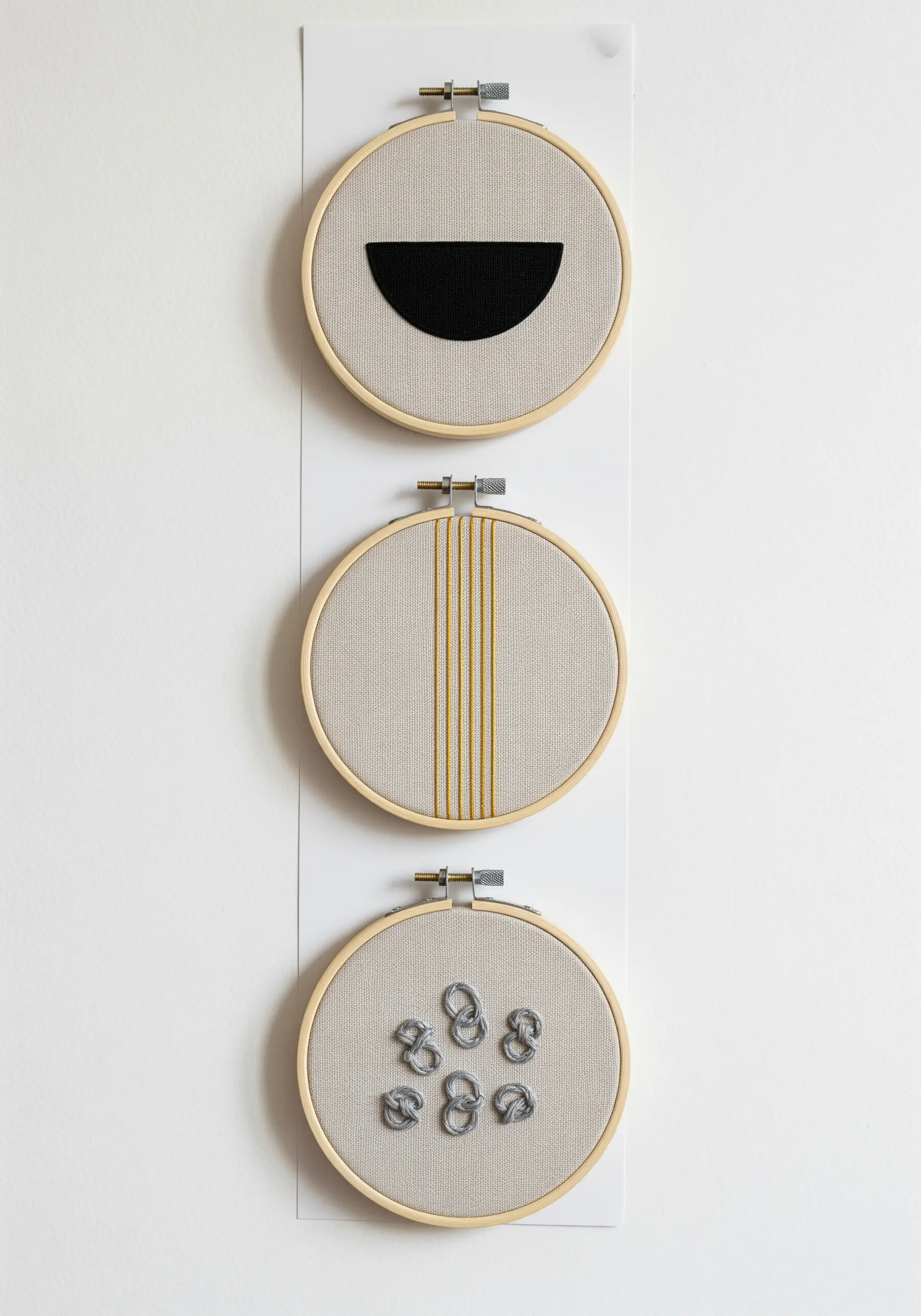 A triptych of three small embroidery hoops, each with a different minimalist abstract design.