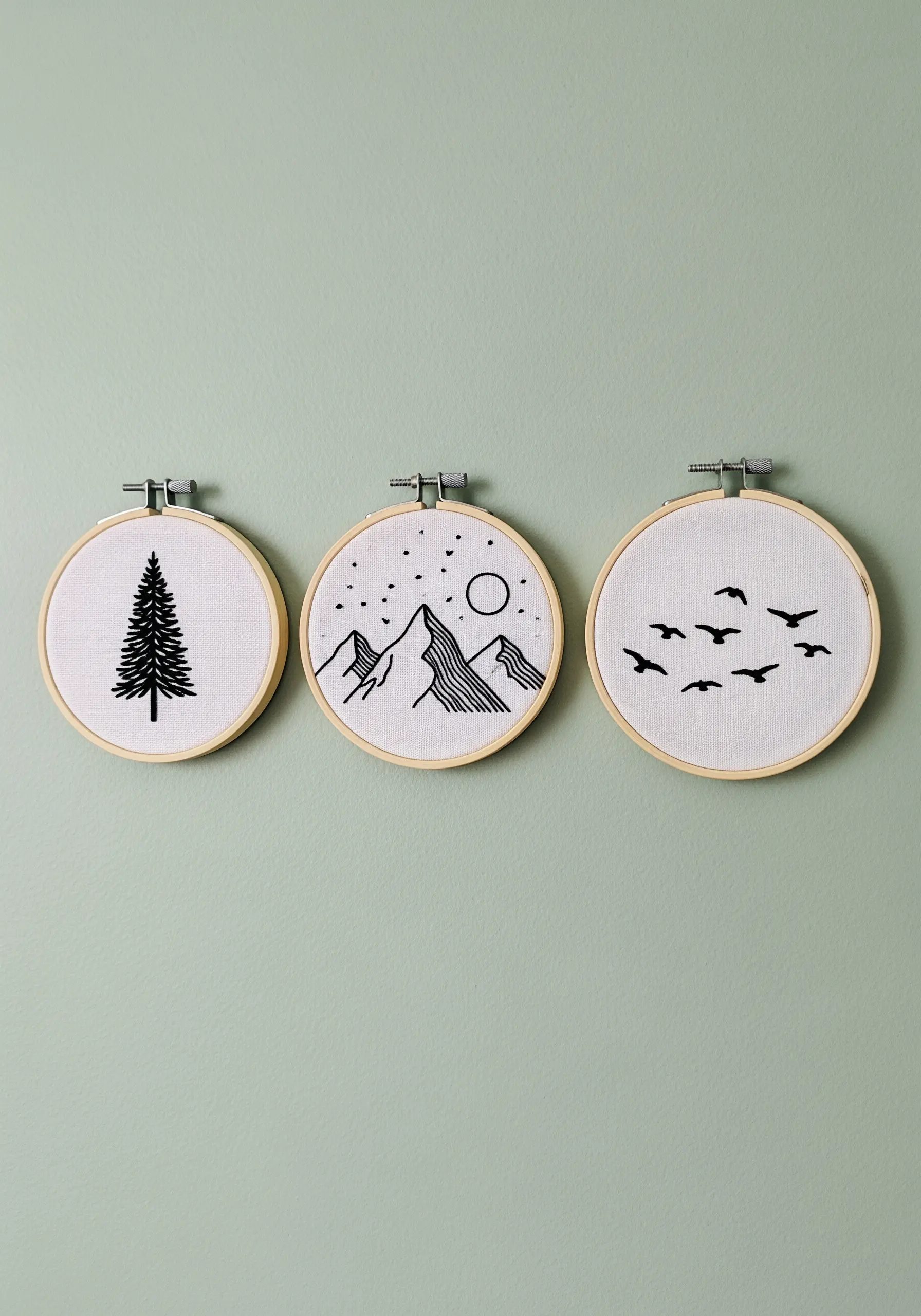 A set of three embroidery hoops with minimalist black-thread designs of a tree, mountains, and birds.