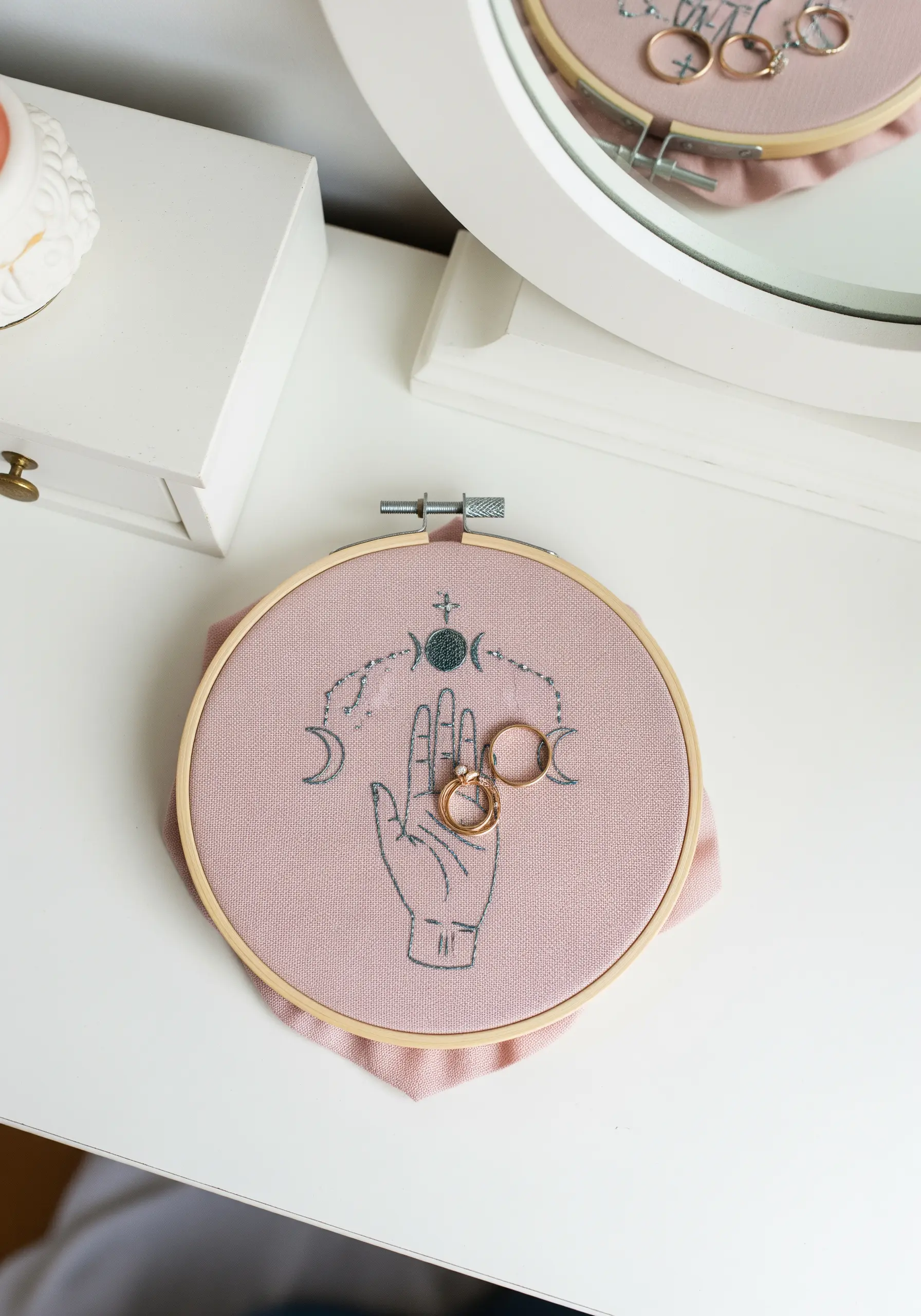 A pink embroidery hoop with a mystical hand and moon design, used to hold rings.