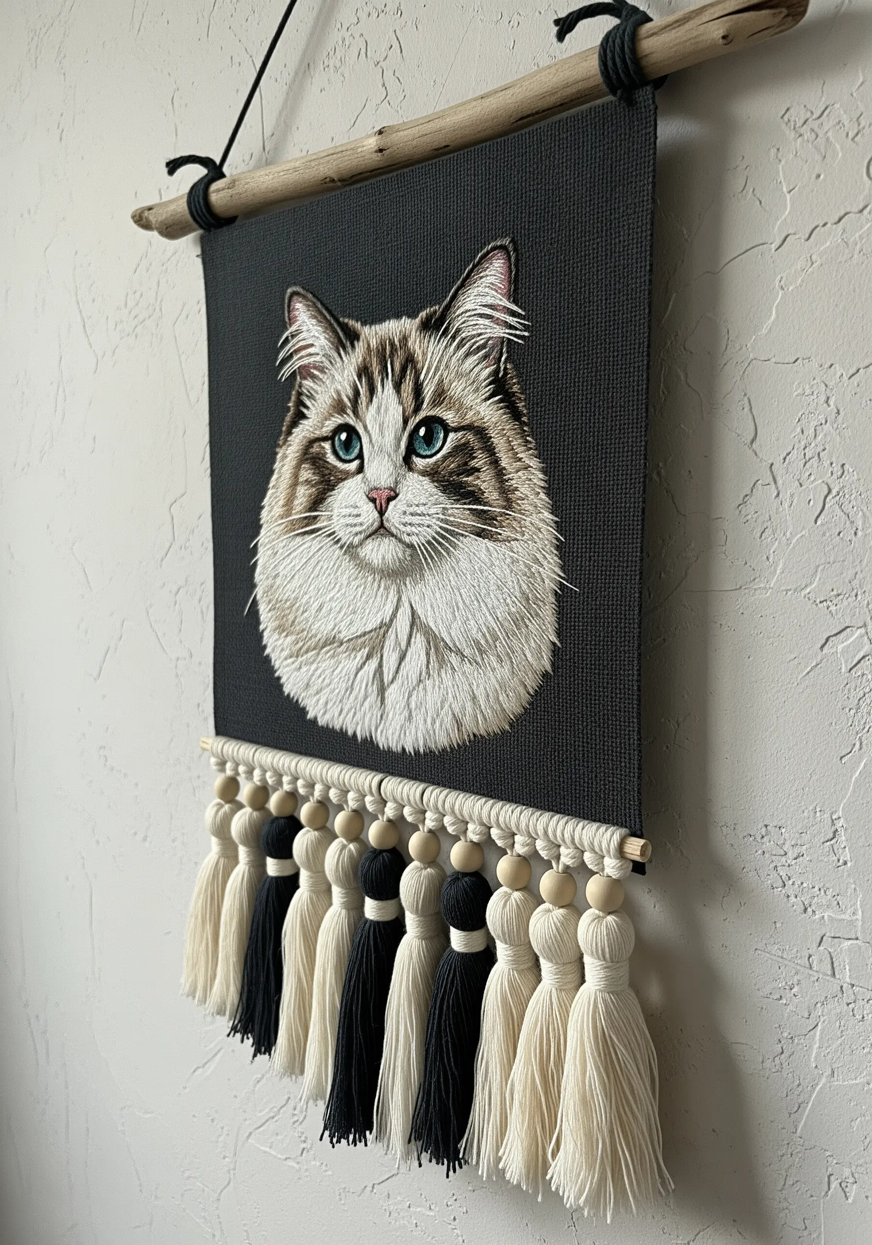 A realistic cat portrait embroidered on dark fabric, finished as a wall hanging with tassels.