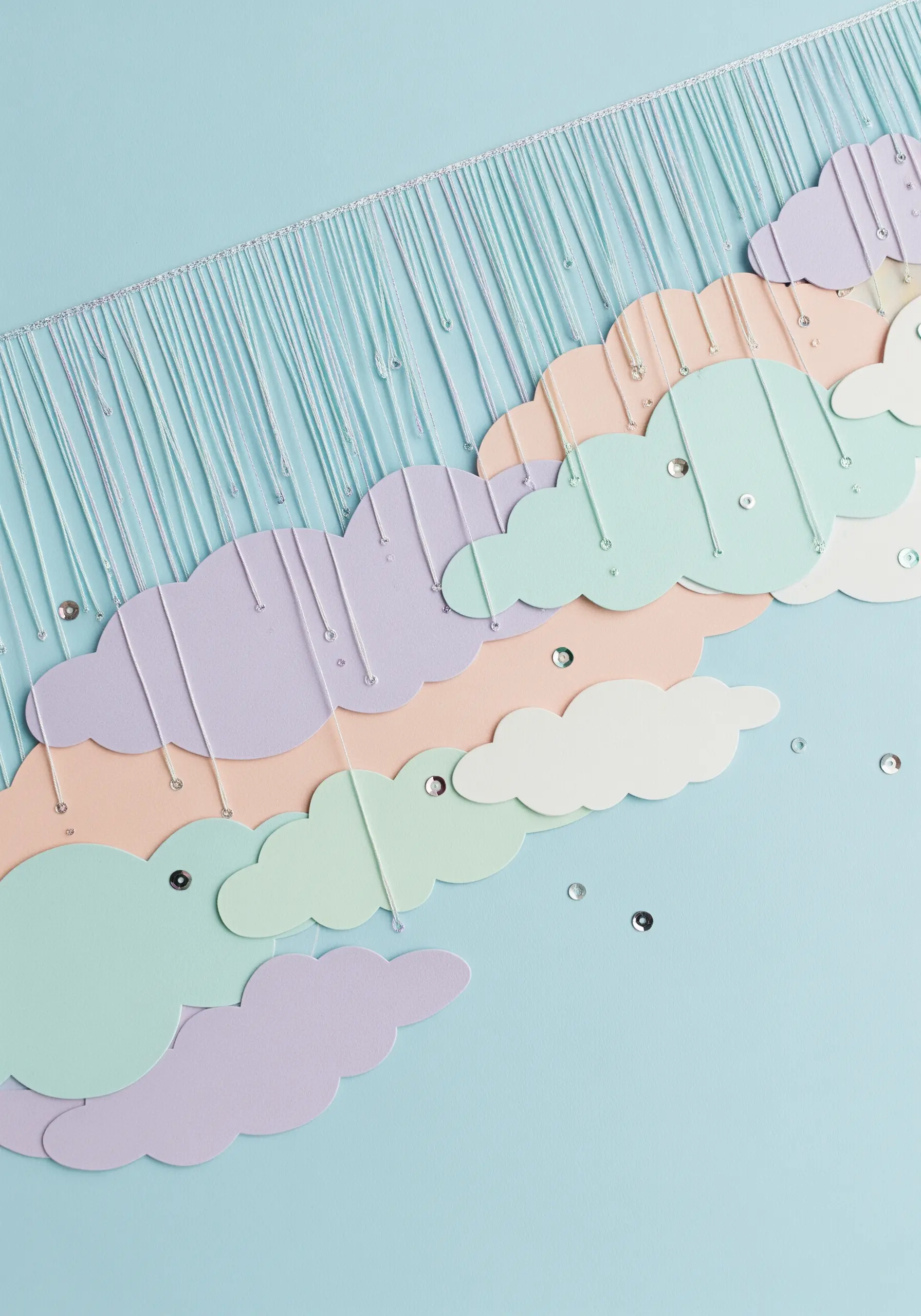 A paper collage of pastel clouds with threads hanging down like rain, embellished with tiny sequins.