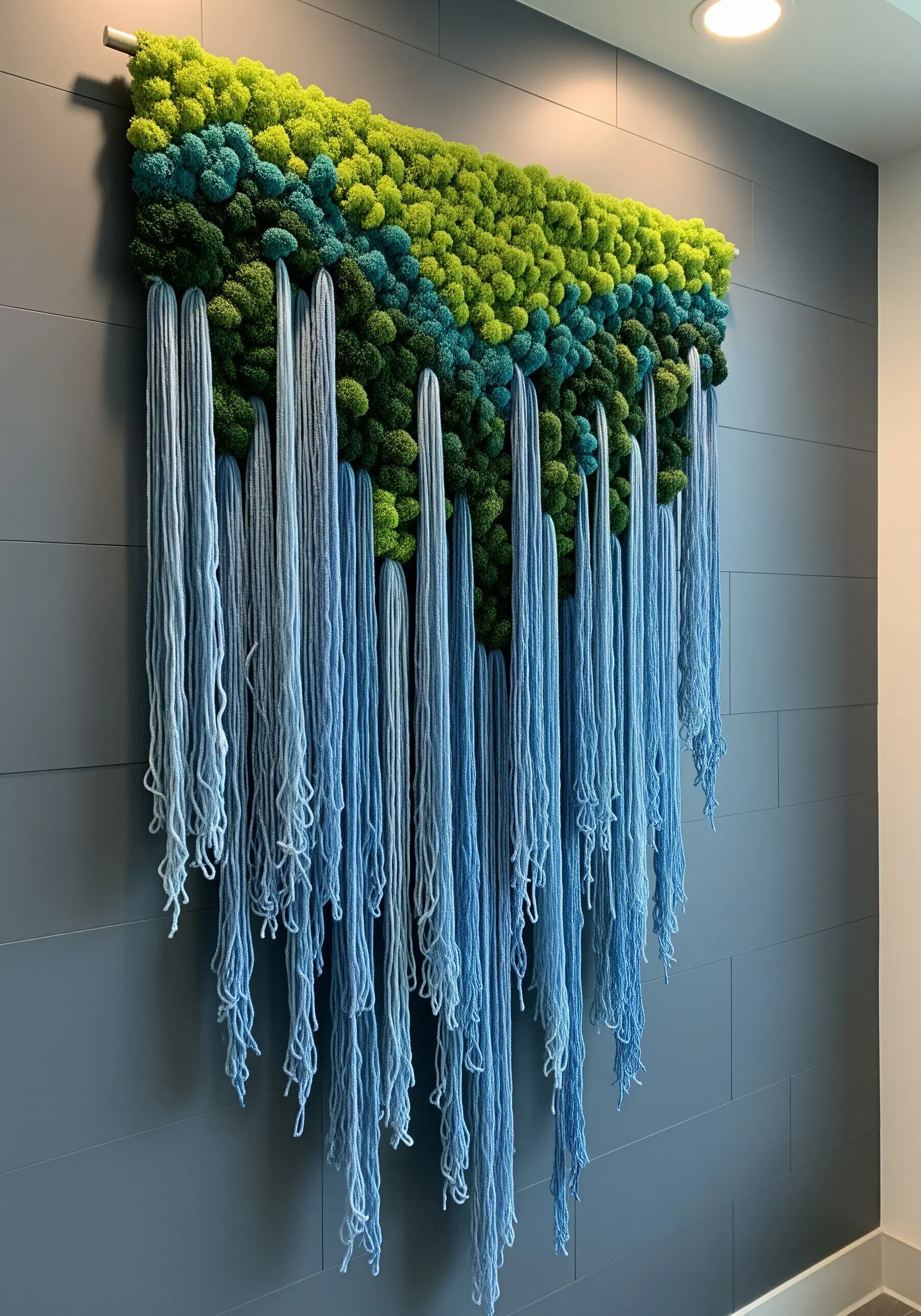 A textured wall hanging with a green moss top and long, flowing blue yarn creating a waterfall effect.