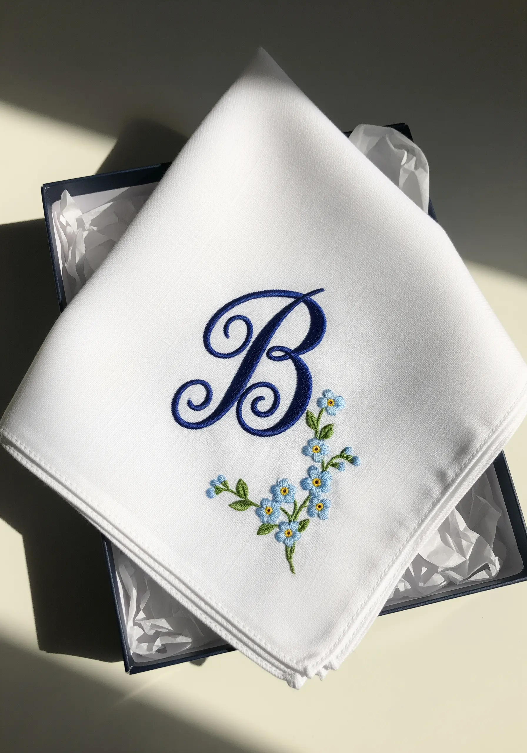A white handkerchief with a beautifully embroidered navy blue 'B' monogram and tiny blue flowers.