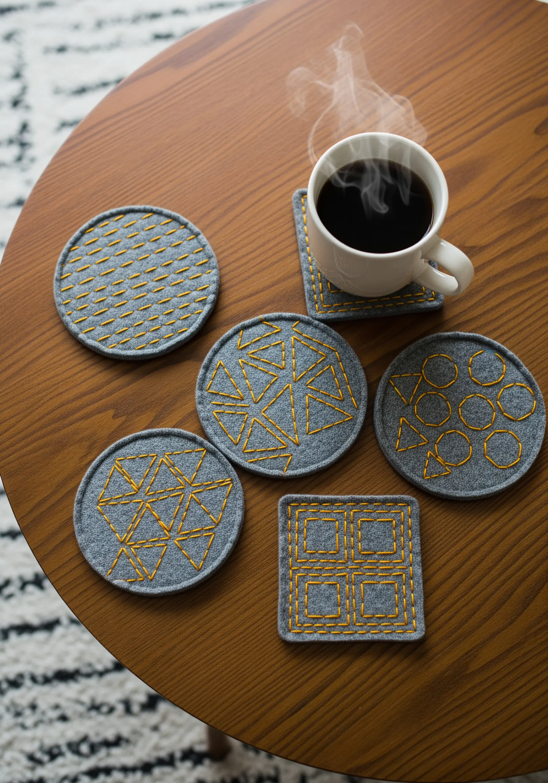 A set of gray felt coasters with modern geometric patterns stitched in yellow thread.