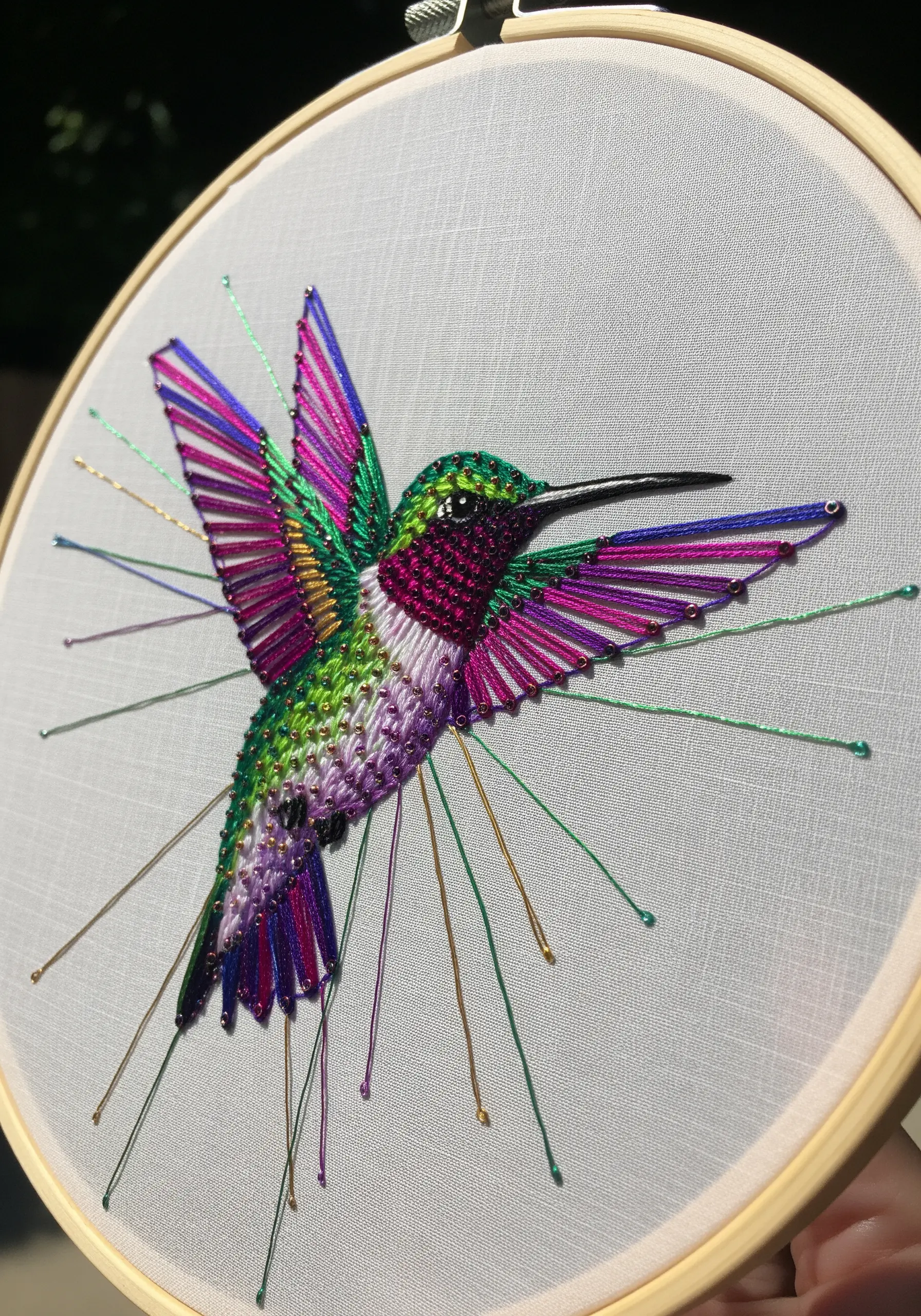 Embroidered hummingbird with iridescent threads and stitches extending beyond the wing outline.