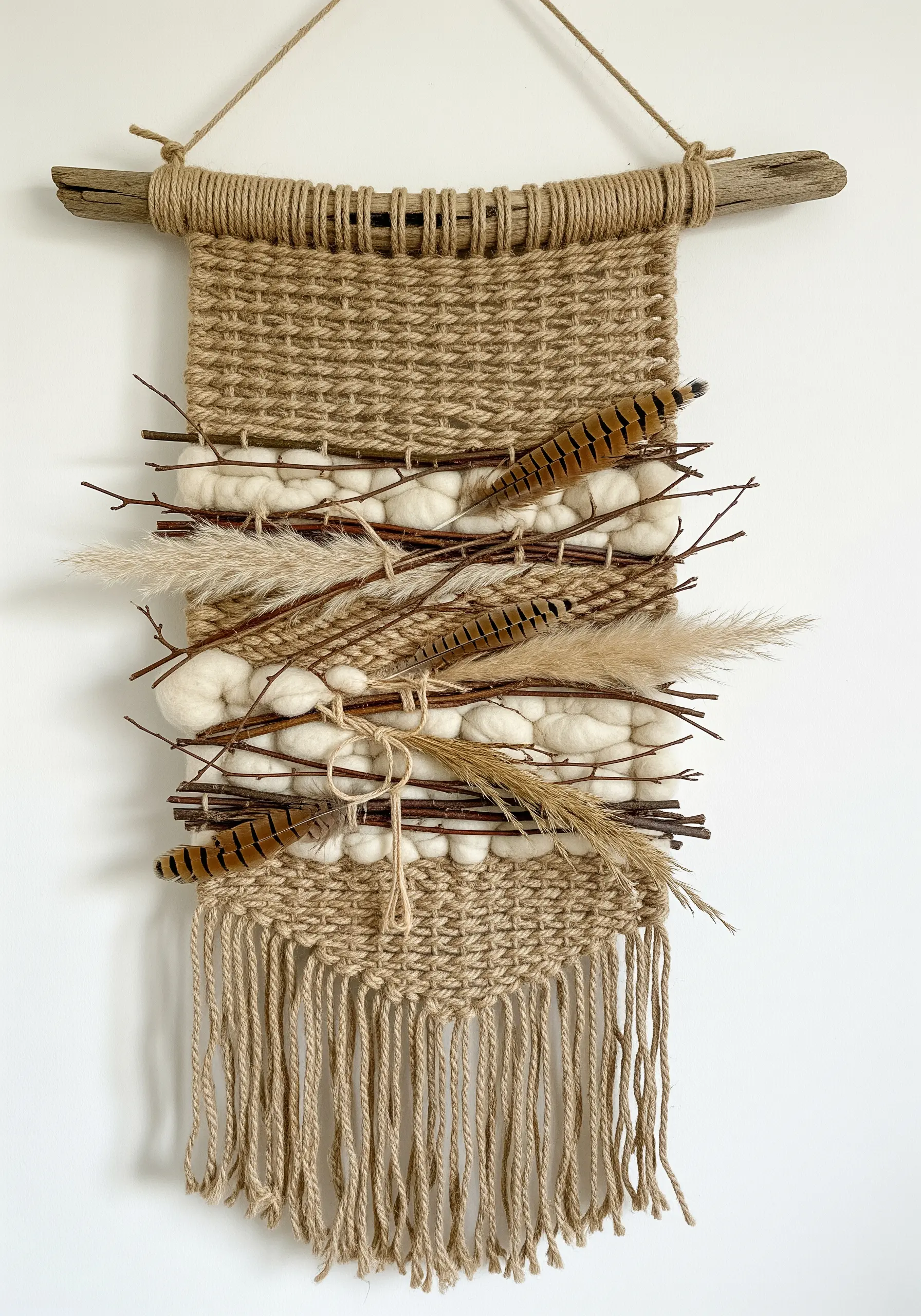 A rustic woven wall hanging made of jute, incorporating twigs, feathers, and dried grass.